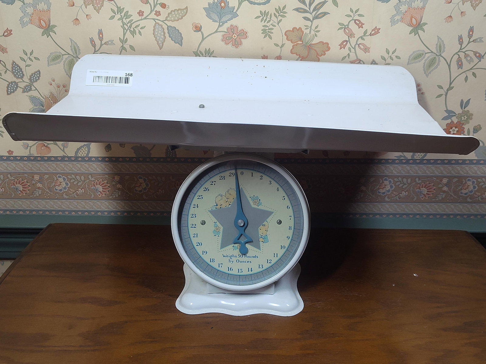 Vintage Mechanical Nursery Baby Scale: This vintage mechanical nursery scale is composed of sheet metal with a white enamel finish. It features a pedestal base supporting a large, curved weighing tray designed for infant use. The circular