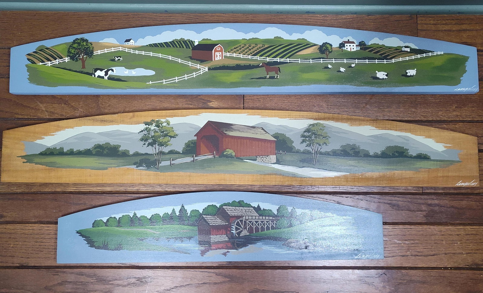 Langelier: Trio of Americana Folk Art Landscapes: A trio of contemporary Americana folk art landscape paintings executed in acrylic on shaped wood panels. Each panel is designed with a gently arched top and features a distinct pastoral scene rendered