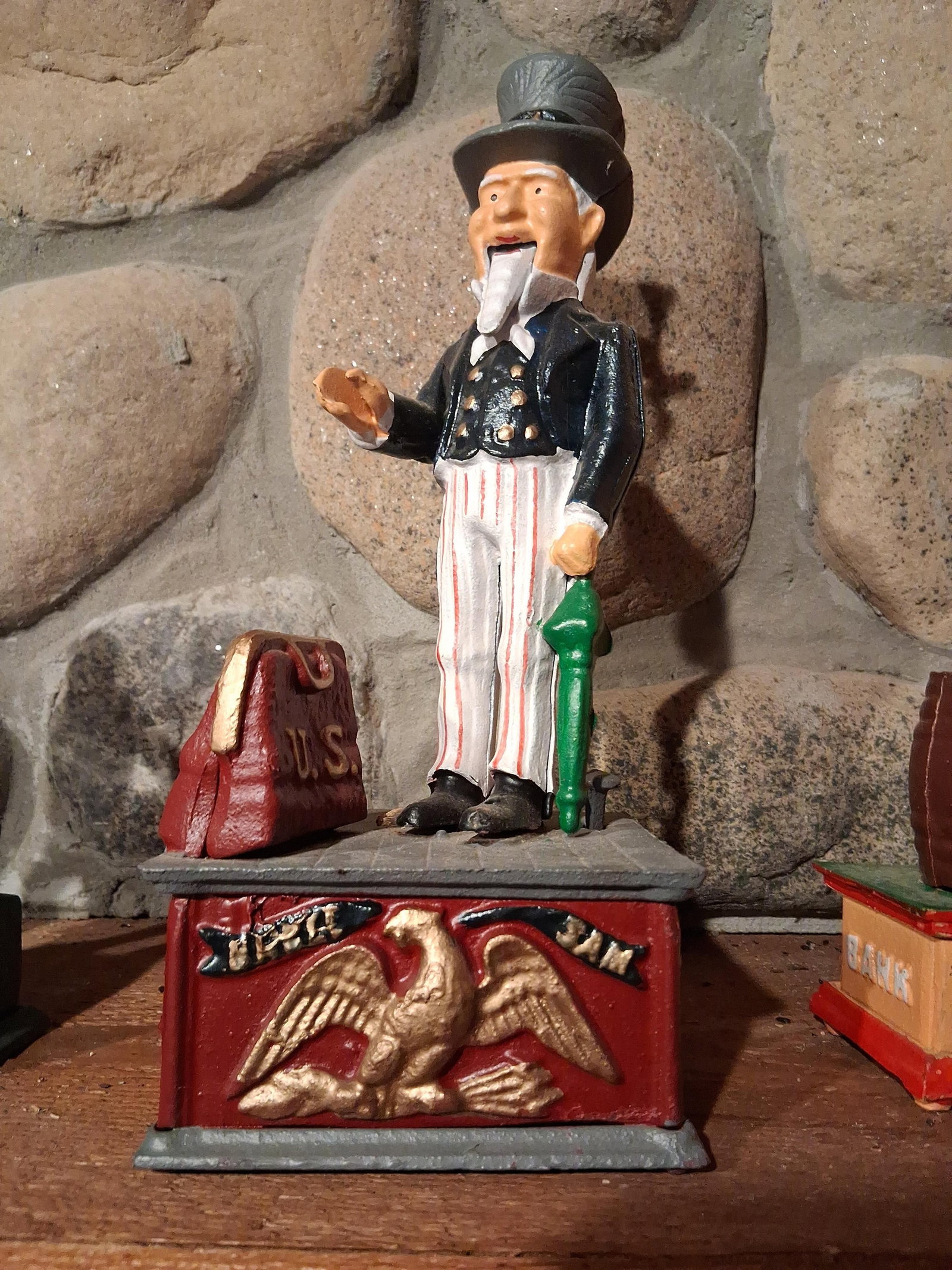 Vintage Uncle Sam Mechanical Bank: This mechanical coin bank is a vintage reproduction of the 1886 design by Shepard Hardware Co. The figure represents Uncle Sam standing on a grey platform next to a red carpet bag. The front of the