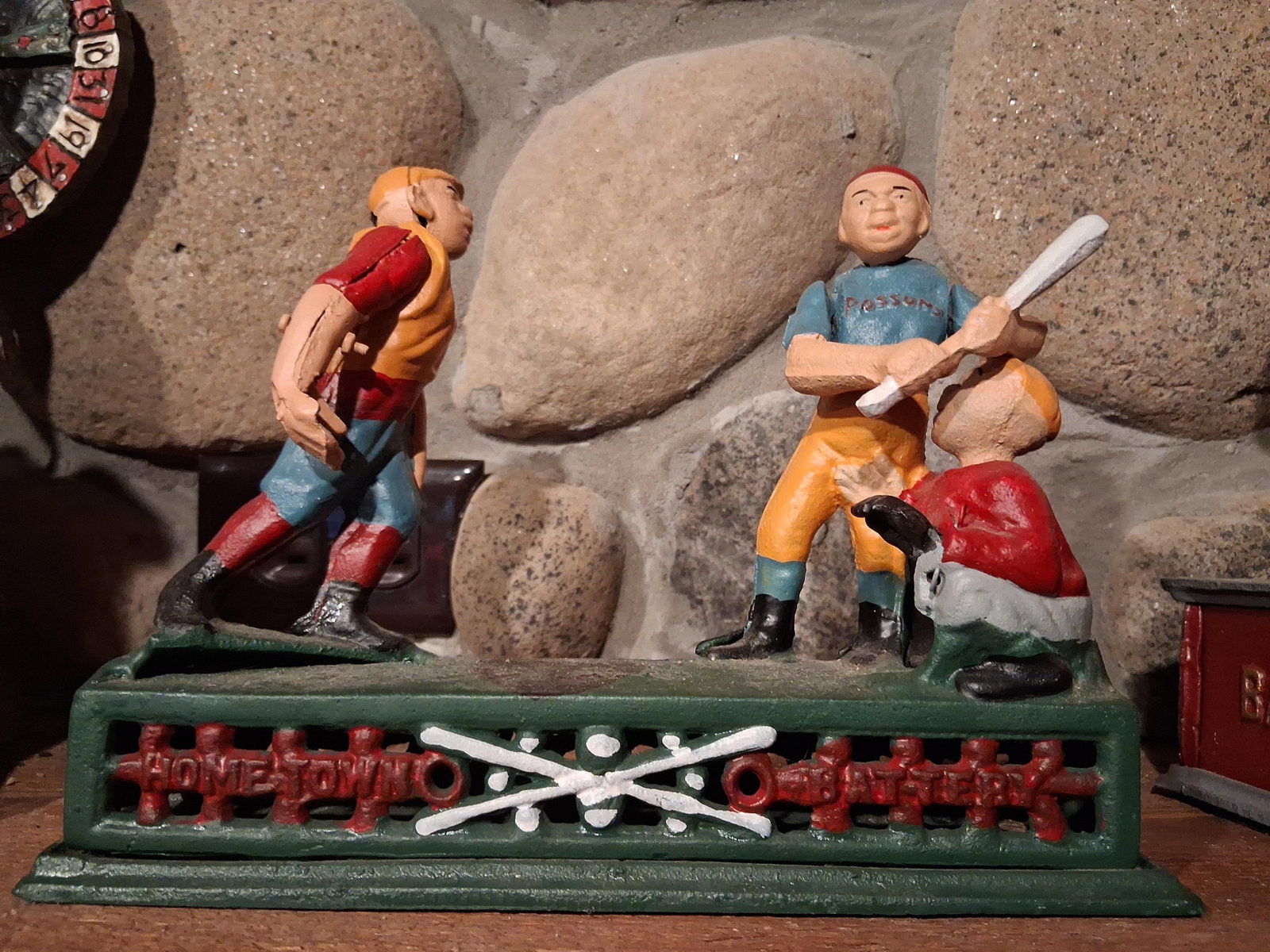 Hometown Battery Mechanical Baseball Bank: A cast iron mechanical coin bank featuring a baseball theme titled Home Town Battery. The bank depicts three multi colored figures mounted on a rectangular green base: a pitcher, a batter in a blue PO