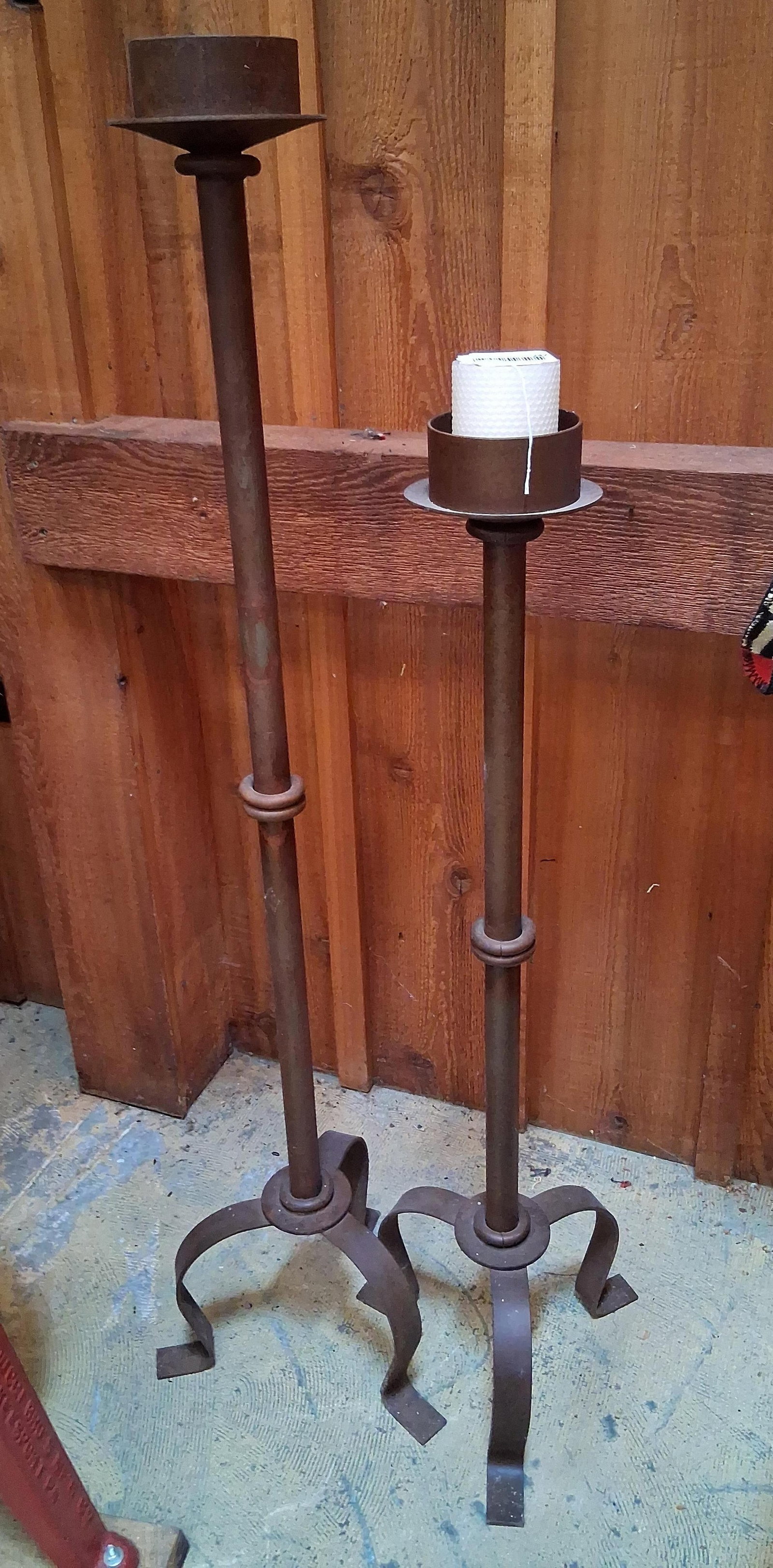 Wrought Iron Candle Stands: A pair of floor-standing candle holders constructed from wrought metal, featuring staggered heights. Each piece consists of a deep, cylindrical candle cup equipped with a central pricket spike