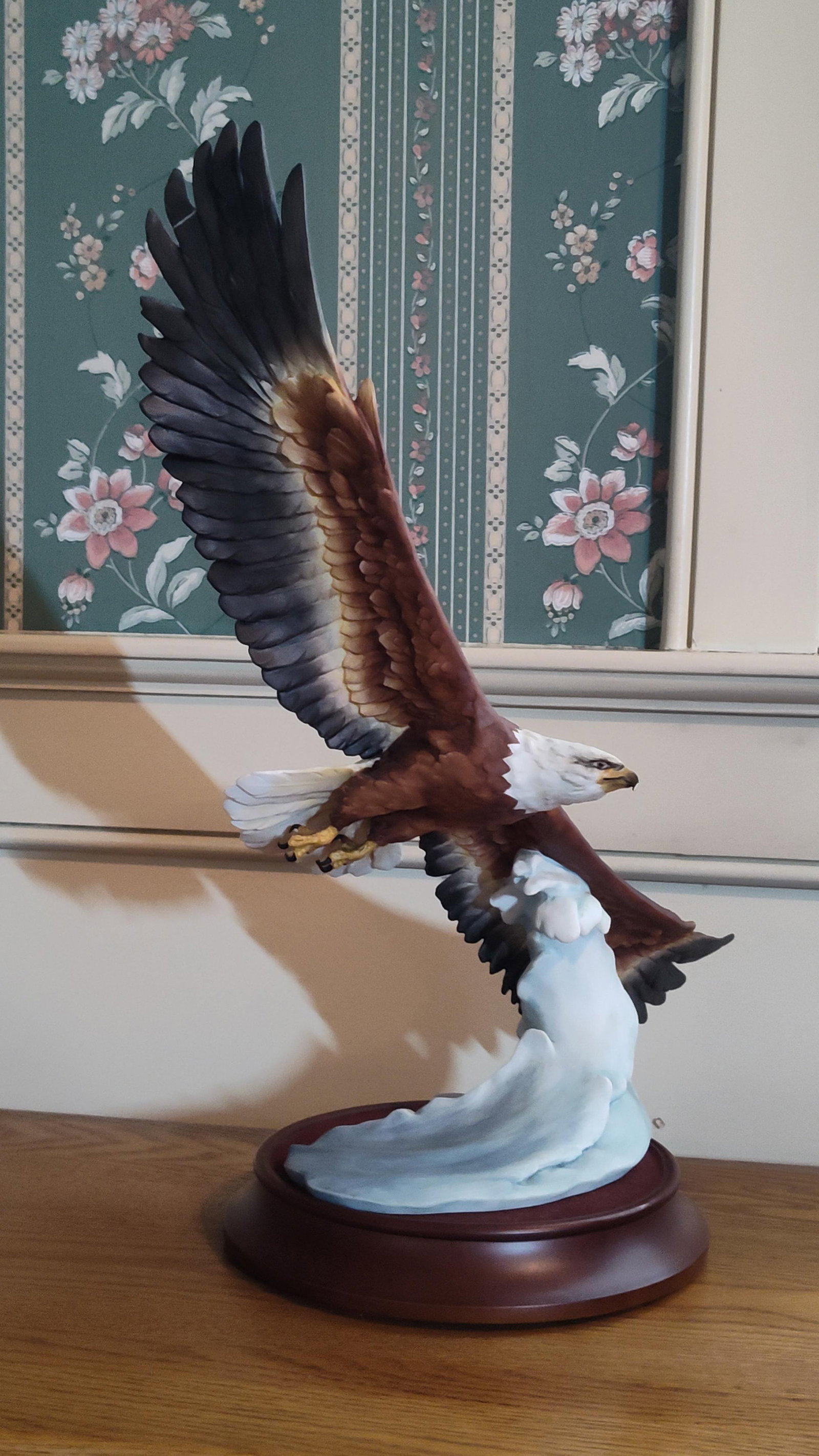 Luqman Gawanika Kaiser Porc. Bald Eagle Figurine: This large-scale wildlife sculpture depicts a bald eagle in flight, banking over a stylized, pale blue and white cresting wave. The piece is constructed of hand-painted bisque porcelain with a matte,
