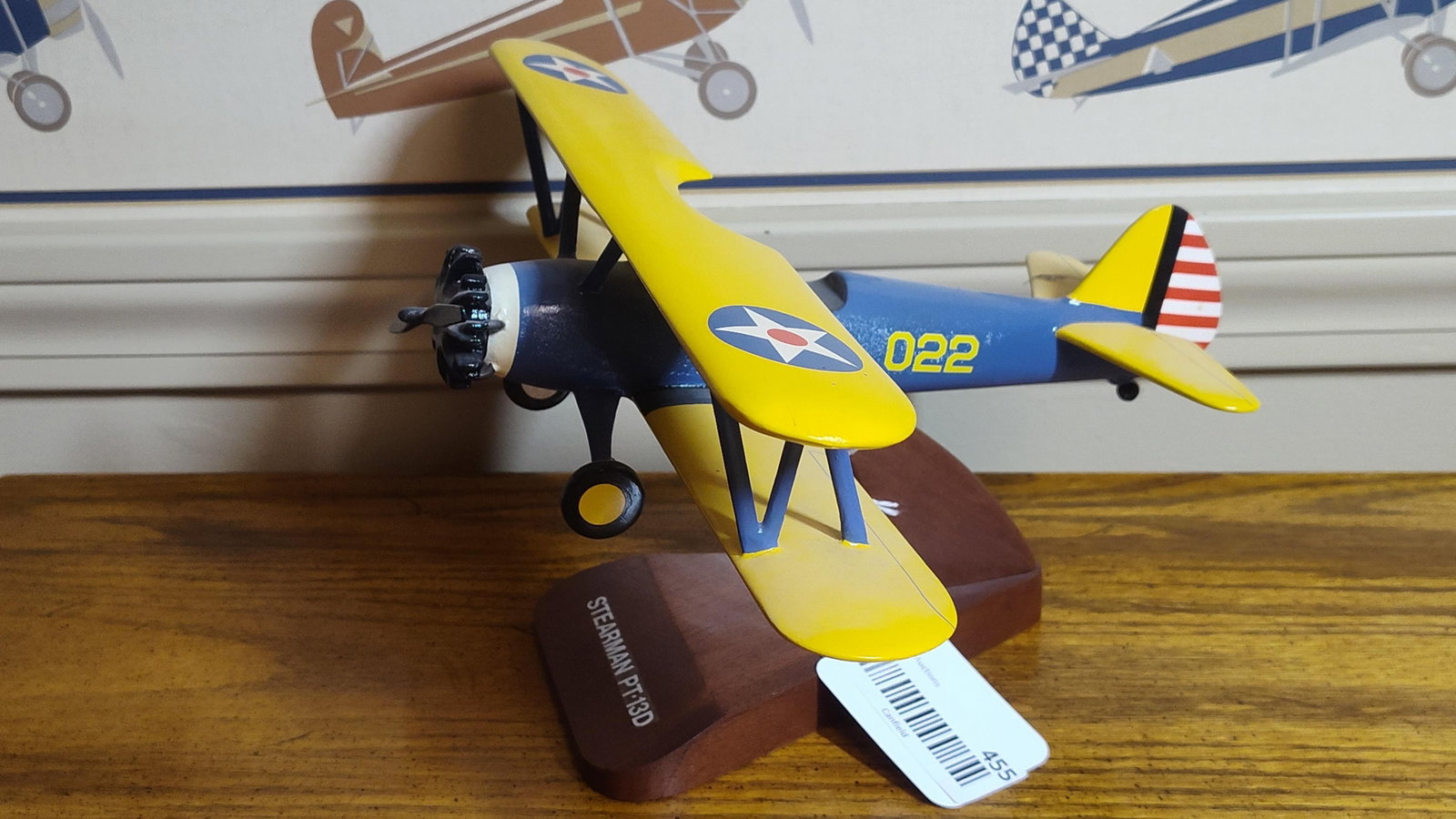 Desktop Display Model Stearman PT-13D Kaydet: A desktop display model of a Stearman PT-13D Kaydet biplane. The item is constructed of hand-painted wood and is mounted on a dark-finished wooden display stand. The aircraft features a traditional U.