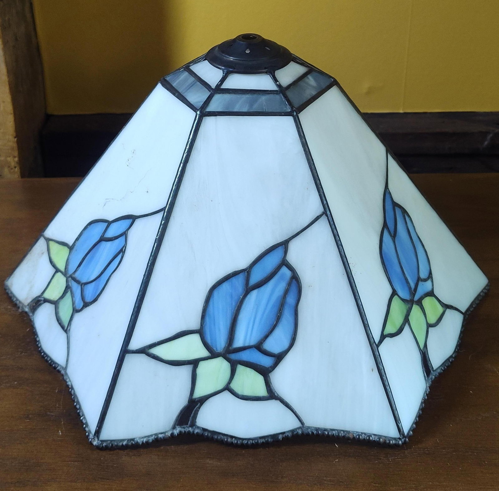 Tiffany Style Slag Glass Floral Lamp Shade: A Tiffany-style leaded slag glass lamp shade featuring a hexagonal, tapered pyramidal form with six panels. Each panel displays a floral motif composed of a blue flower bud with two-toned green leaves