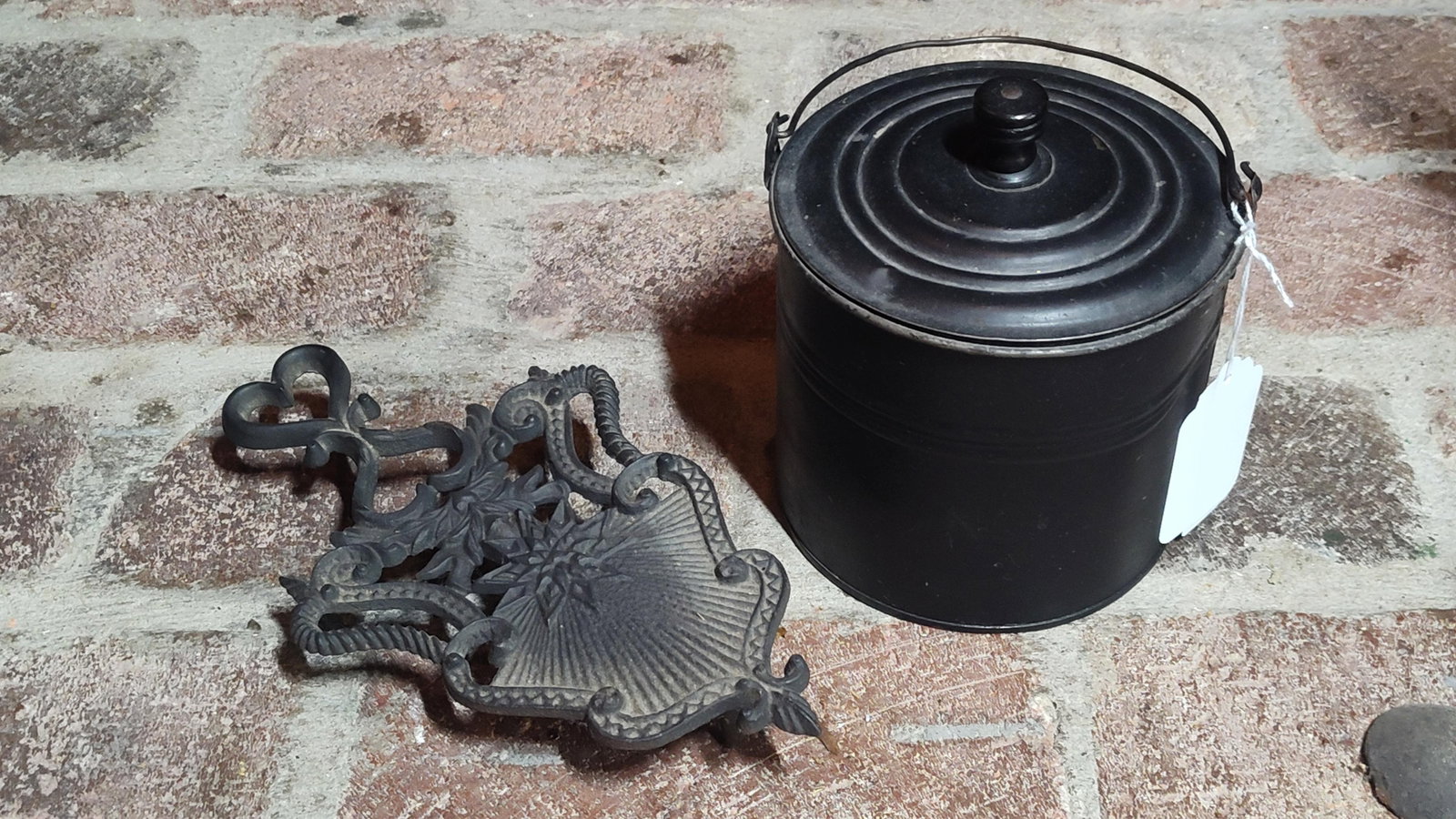Cast Iron Fan Pattern Trivet & Blackened Canister: A lot of two antique metal household items consisting of a cast iron trivet and a cylindrical canister. The trivet features an openwork design with a heart-shaped handle at the top, decorative