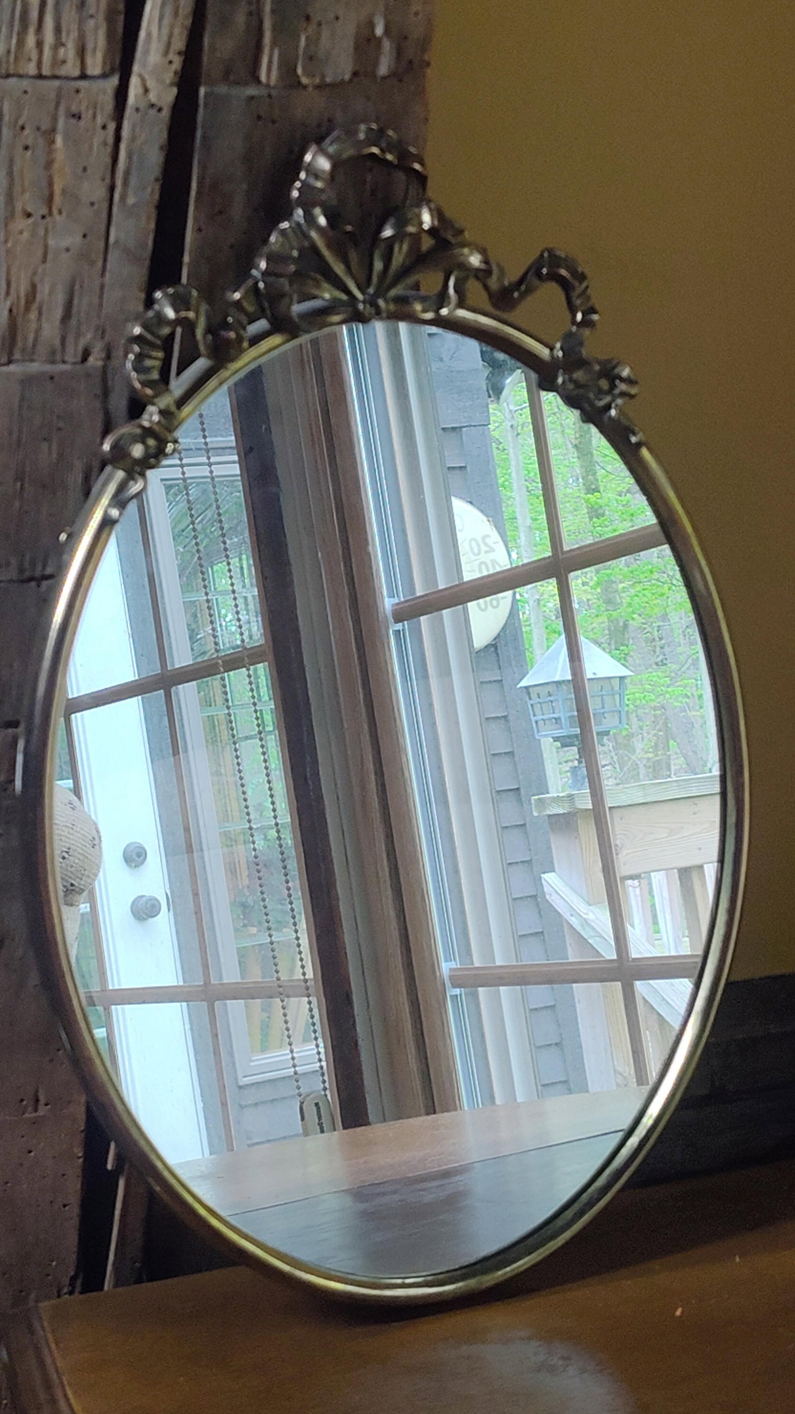 Neoclassical Brass Oval Mirror w/ Ribbon Bow Crest: This vintage wall mirror consists of an oval mirror plate housed in a cast metal frame. The frame, likely composed of brass or a similar copper-based alloy, features a dark golden-brown patina. The de