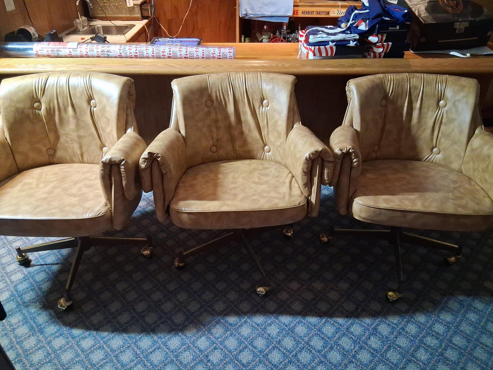 Trio Mid-Century Vinyl Swivel Armchairs on Casters: This set consists of three vintage swivel armchairs in the bucket style, popularized by American manufacturers in the late 1960s and 1970s. The chairs are upholstered in camel-colored vinyl with a mot