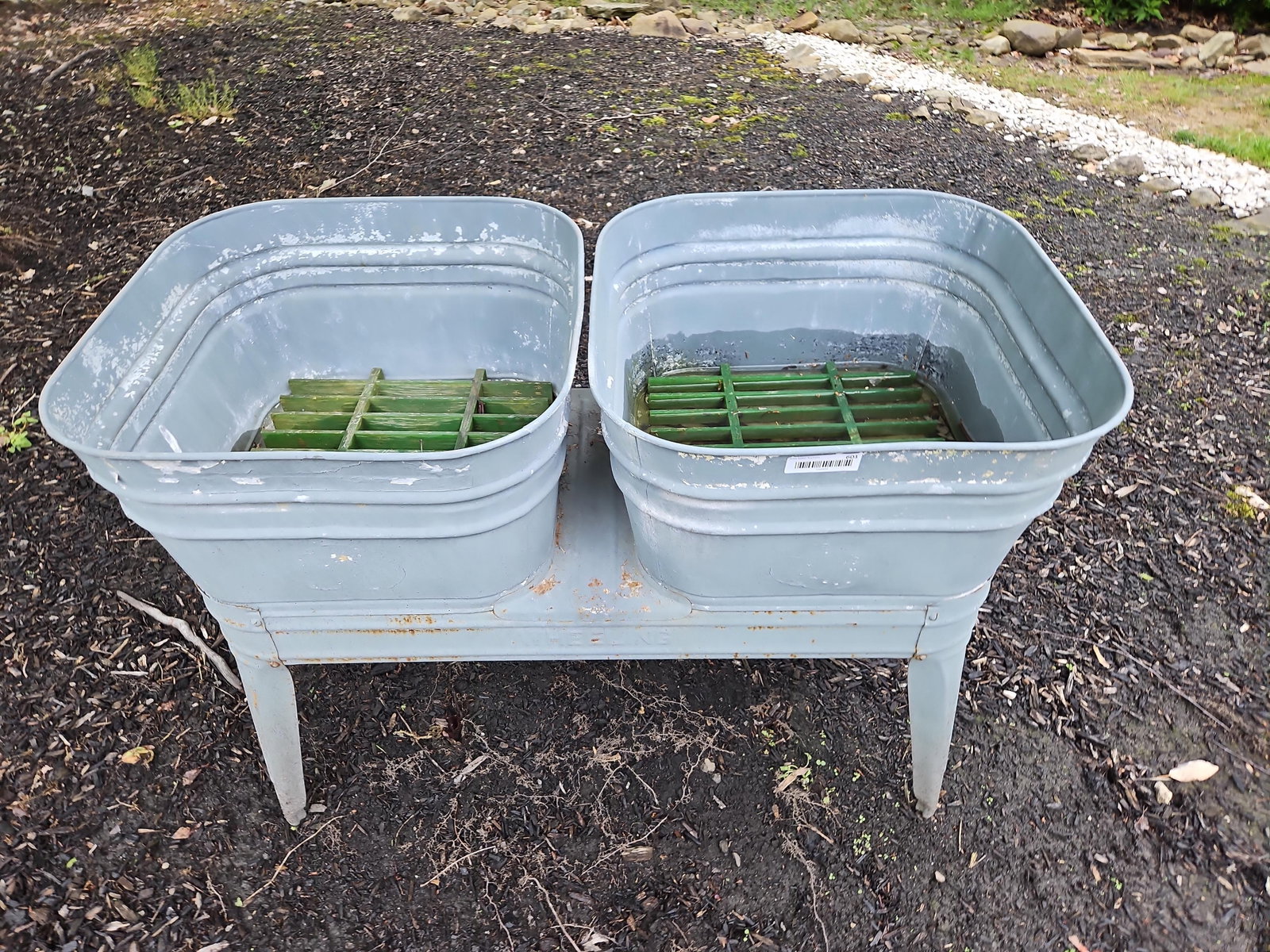 Wheeling Galvanized Double Wash Tub on Stand: Manufactured by the Wheeling Corrugating Company, this vintage double-basin laundry tub is constructed from galvanized steel. The unit features two rectangular basins joined by a central bridge and se