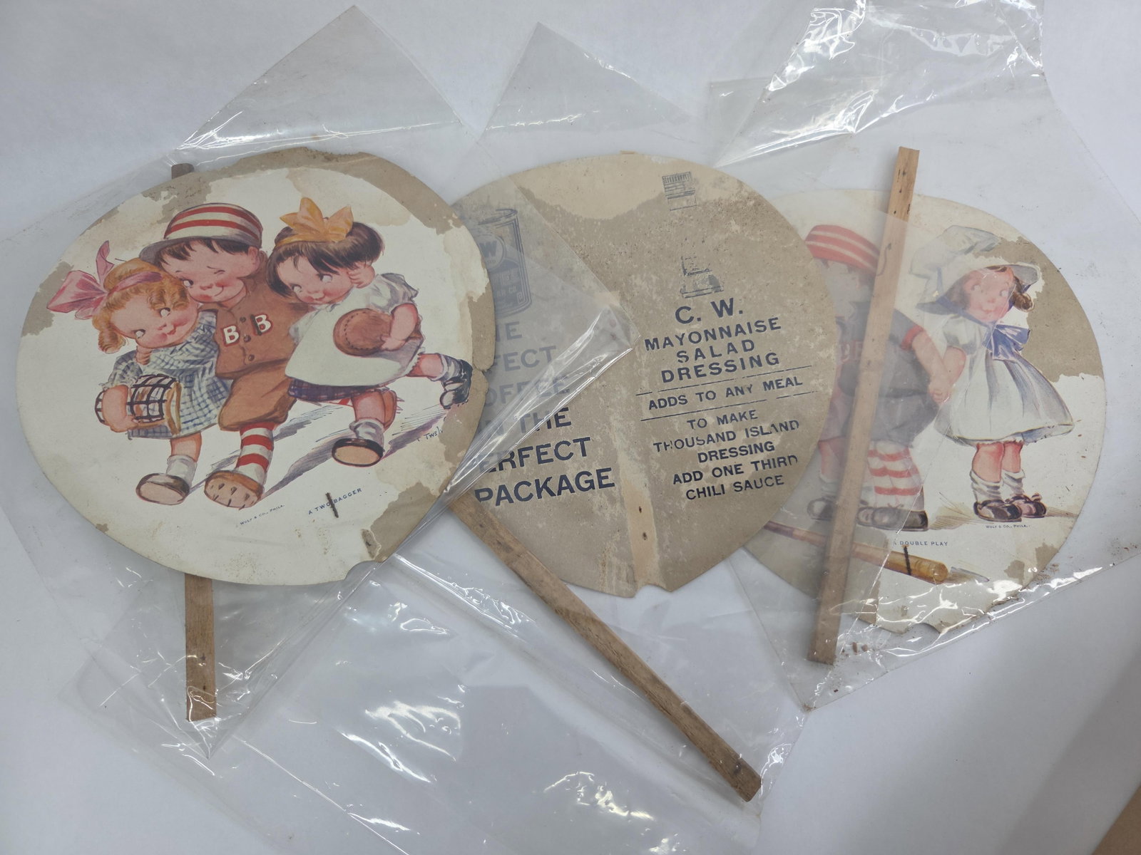 A Trio of Antique Charles Twelvetrees Baseball-Themed C.W. Brand Advertising Hand Fans: A trio of antique cardboard advertising hand fans dating to the early 20th century, approximately 1915 to 1925. The front of each fan features a color lithograph illustration of children in