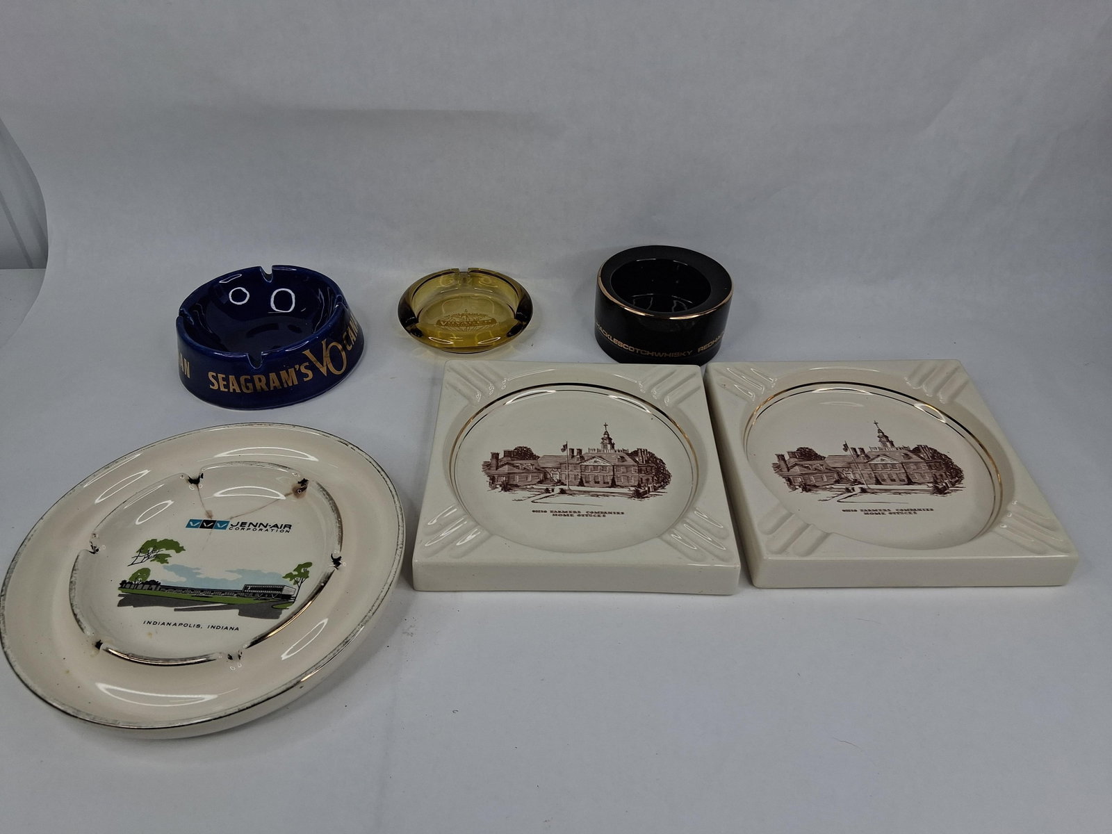 Set 6 Advertising Ashtrays: Seagrams VO & Ohio: This collection consists of six advertising ashtrays manufactured from various materials including glazed ceramic, earthenware, and pressed glass. The lot includes a matching pair of square off-white