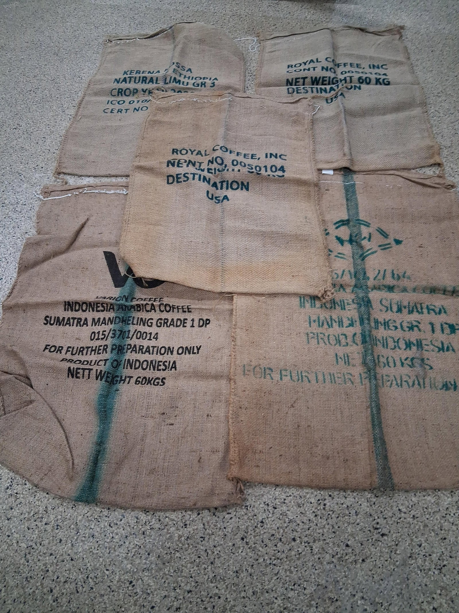 A Collection of Five Burlap Coffee Transport Sacks: A collection of five woven jute burlap coffee transport sacks originating from Ethiopia and Indonesia. One sack features dark blue text identifying the contents as Kebena Kossa Ethiopian coffee, speci