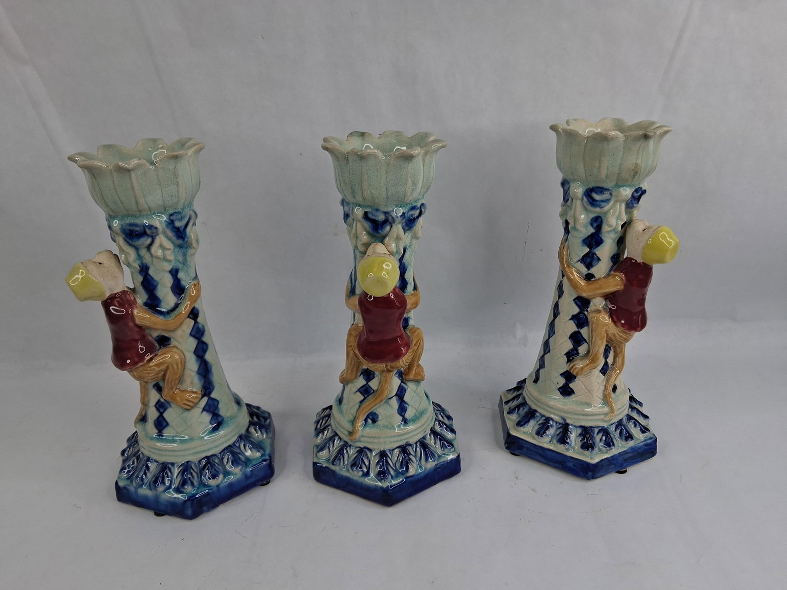 Trio Italian Majolica Figural Monkey Candlesticks: This trio of figural ceramic candlesticks is crafted in the majolica style and originates from Italy. Each piece features a tapered, columnar shaft resting on a stepped hexagonal base and is topped