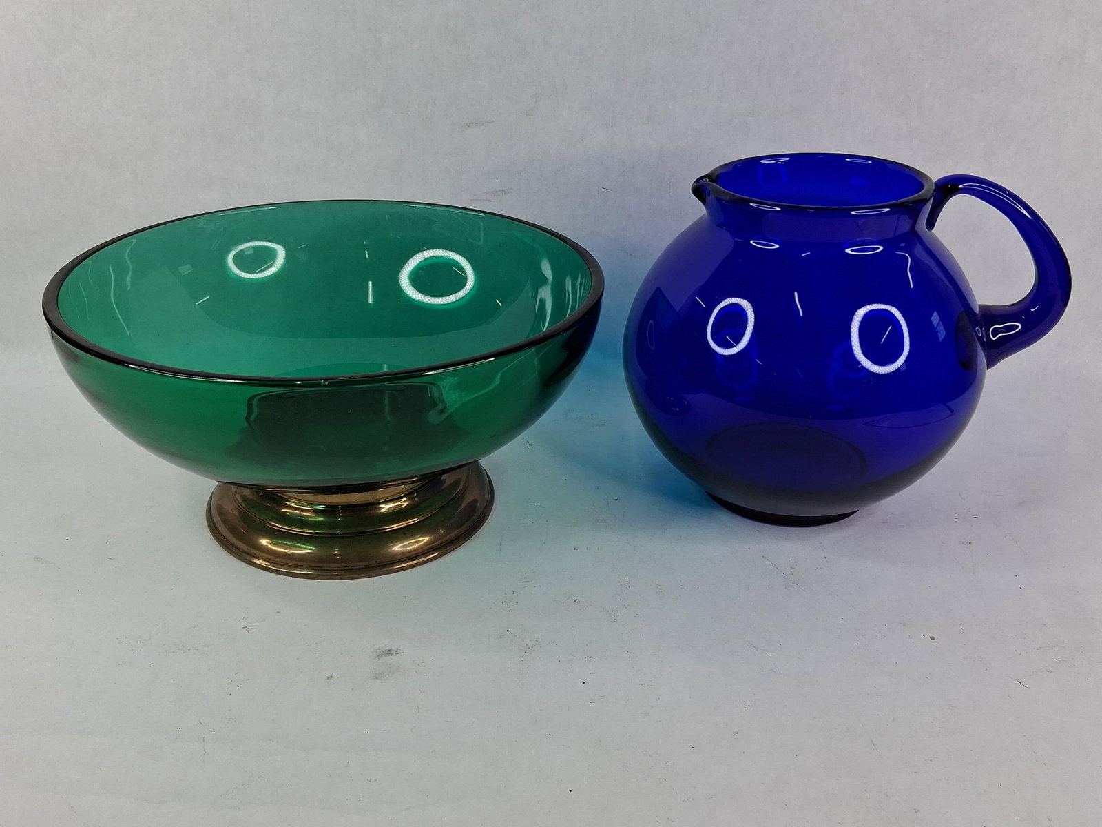 Two Pieces of Mid-Century Modern Art Glass including a Pedestal Bowl and Cobalt Blue Pitcher: This lot features two items of mid-century modern decorative glassware. The first is a deep, round bowl of transparent emerald green glass with a polished rim, set into a tiered brass-tone metal