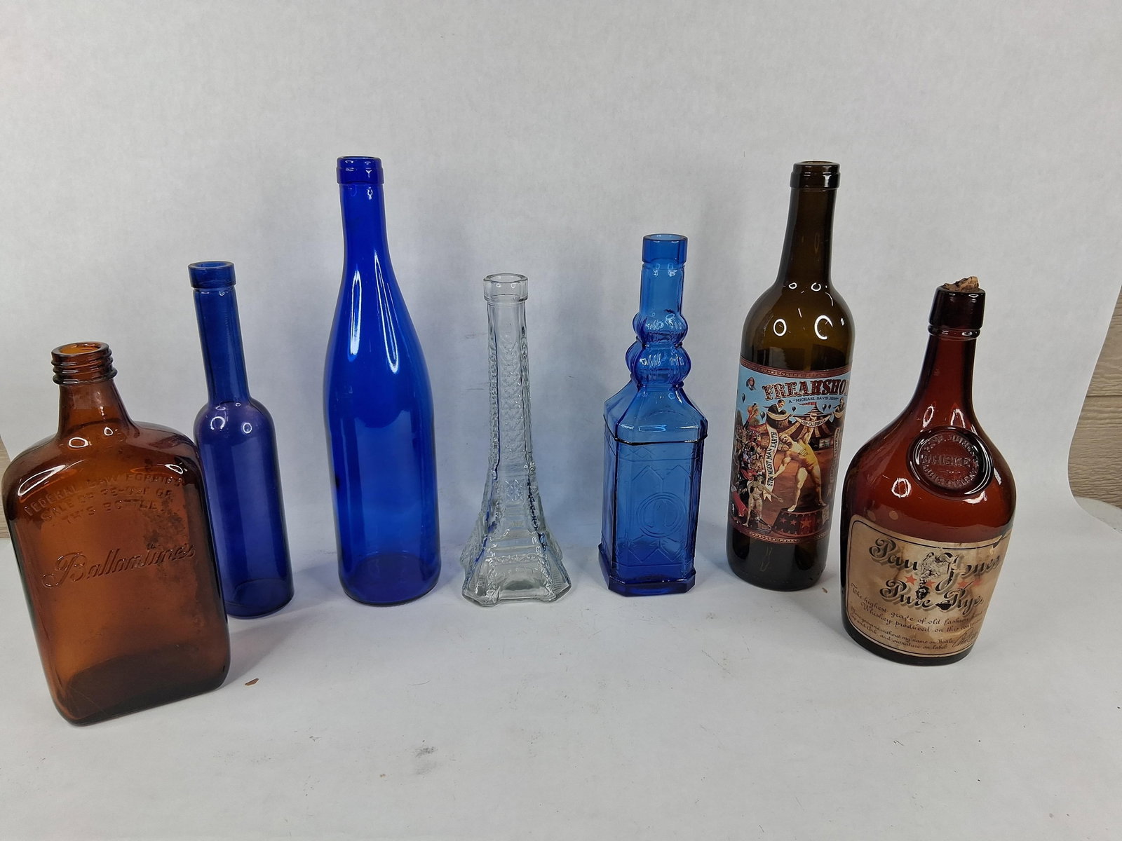 Set Advertising Whiskey & Wine Glass Bottles: An assortment of glass bottles in various shapes, sizes, and colors, including cobalt blue, amber, olive, and clear glass. The collection features a variety of commercial and decorative designs. One a