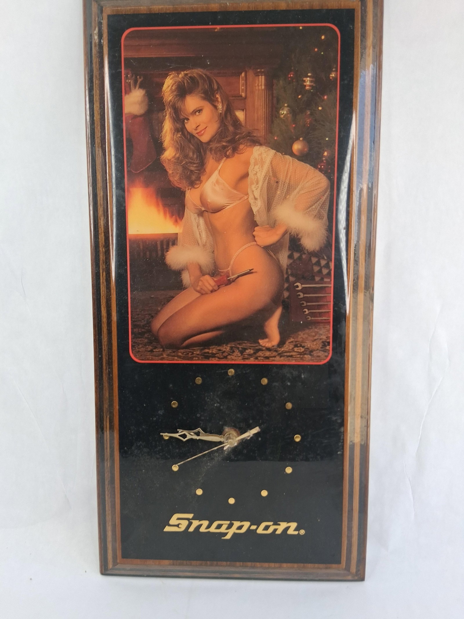 Snap-on Pinup Girl Christmas Wall Clock Plaque: 11x23 A promotional wall clock produced for Snap-on Tools. The item consists of a rectangular wood-grain plaque featuring a color photograph of a pin-up model in a holiday setting. The model is