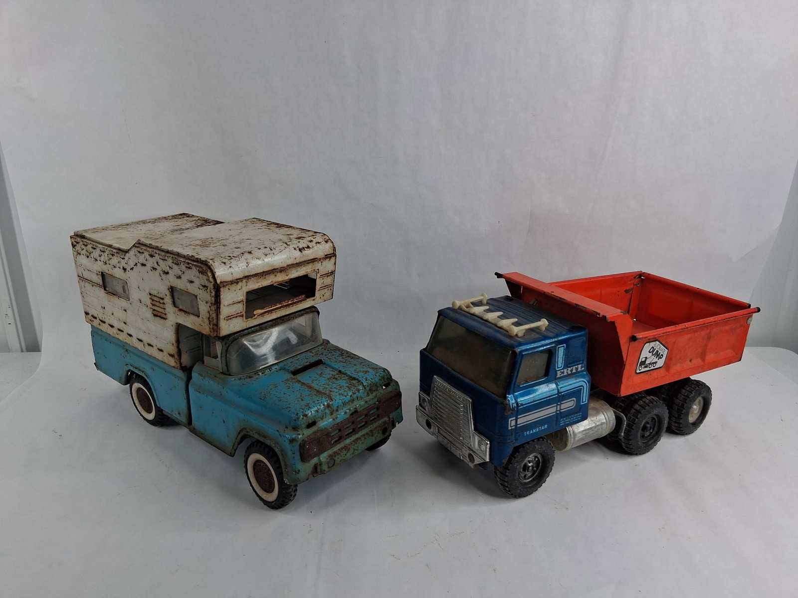 Buddy L Camper & Ertl Transtar Dump Steel Trucks: This lot consists of two toy trucks constructed primarily of pressed steel. The first item is a blue pickup truck equipped with a white slide-in camper shell. The vehicle features white plastic wheels