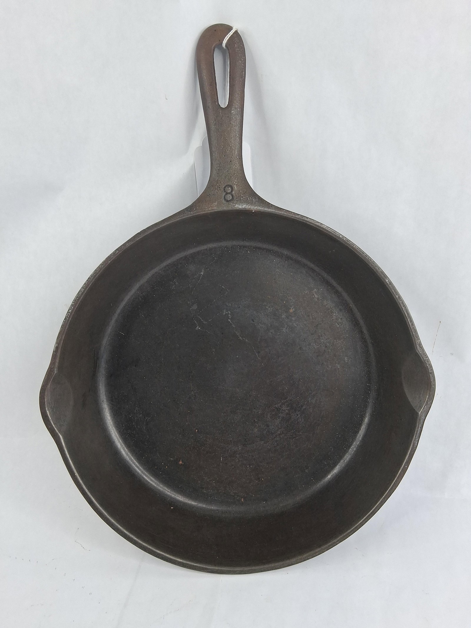 Unmarked No. 8 Cast Iron Skillet: This cast iron skillet, sized as a No. 8, features a traditional round form with two integrated pour spouts and a tapered handle with a teardrop-shaped hanging hole. The upper surface of the handle ne