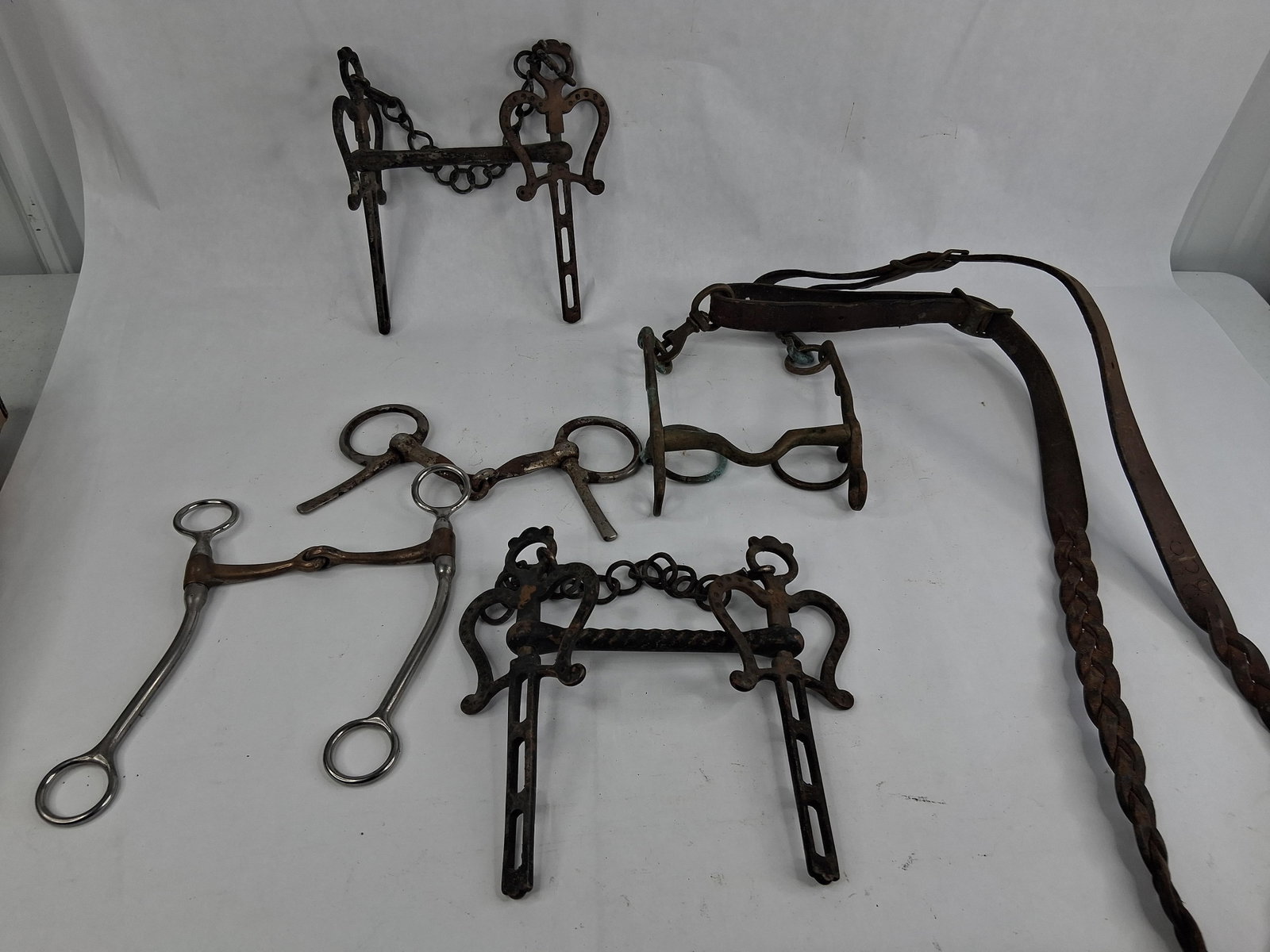 7 Antique & Horse Bits & Braided Leather Rein: This lot consists of a collection of seven items of equestrian tack, including six assorted horse bits and one braided leather rein. The collection includes two ornate curb bits in a traditional