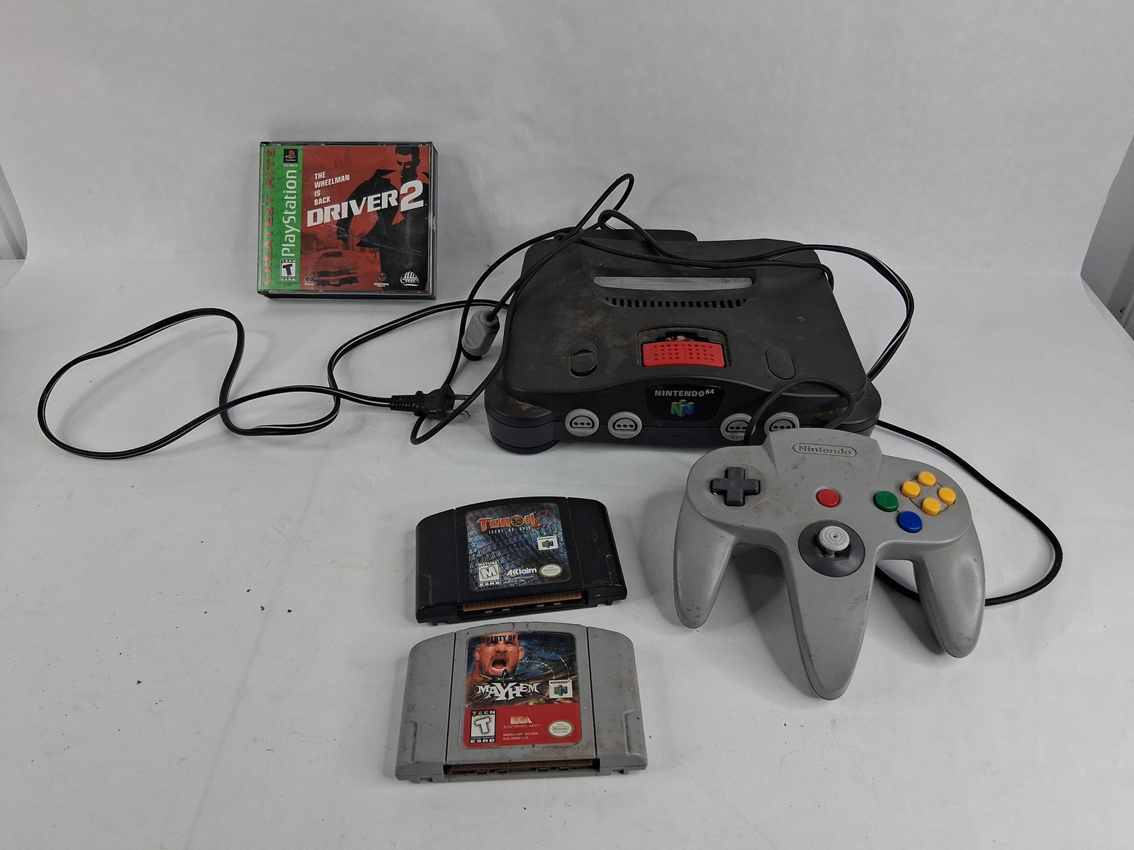 Nintendo 64 & PlayStation Gaming Equipment: A collection of video game electronics featuring a Nintendo 64 Control Deck, model NUS-001(USA). The console is constructed from charcoal gray plastic and is fitted with a red Expansion Pak in the