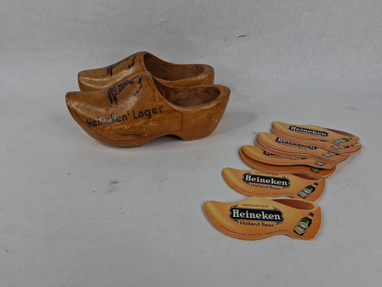 Pair Heineken Wooden Clogs & Paper Coasters: This lot features a pair of miniature wooden clogs and a collection of clog-shaped promotional paper booklets related to Heineken Holland Beer. The wooden clogs are constructed from carved wood with