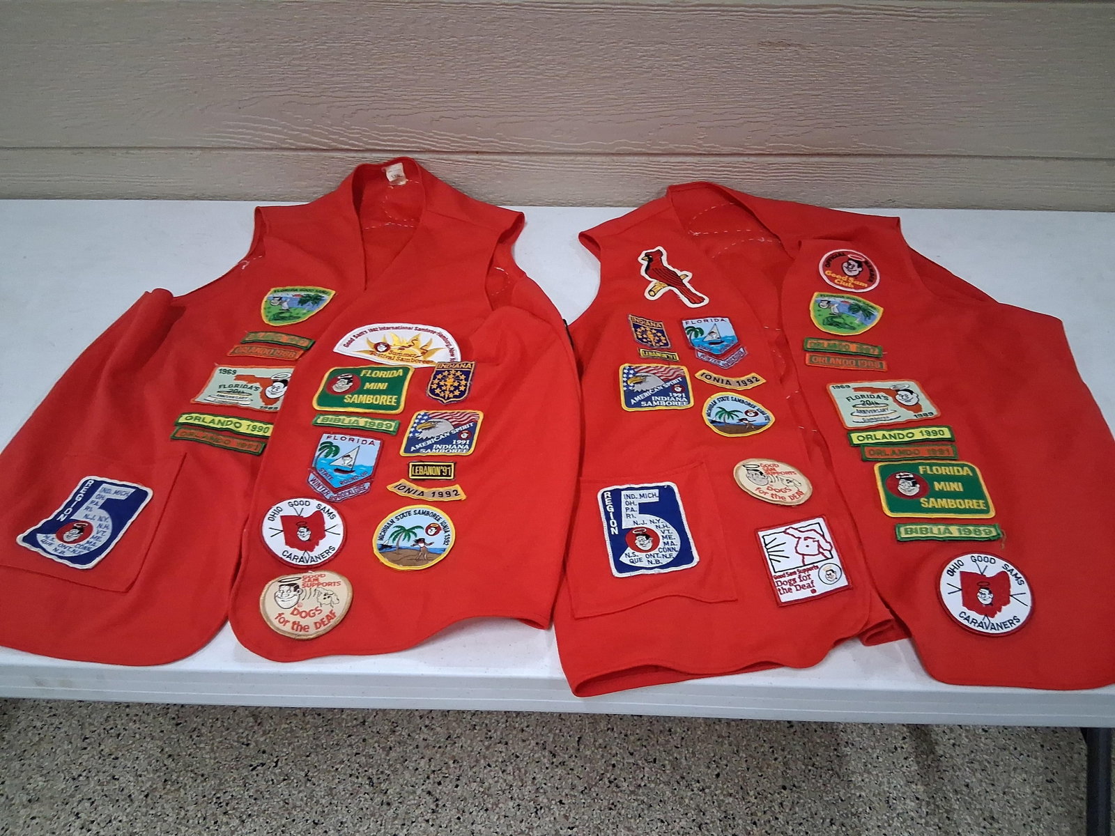 Pair Good Sam Club Red Vests w/ Patches: A pair of red textile vests heavily decorated with a variety of embroidered patches and rockers related to the Good Sam Club international RV organization. The vests are made of a red fabric, likely