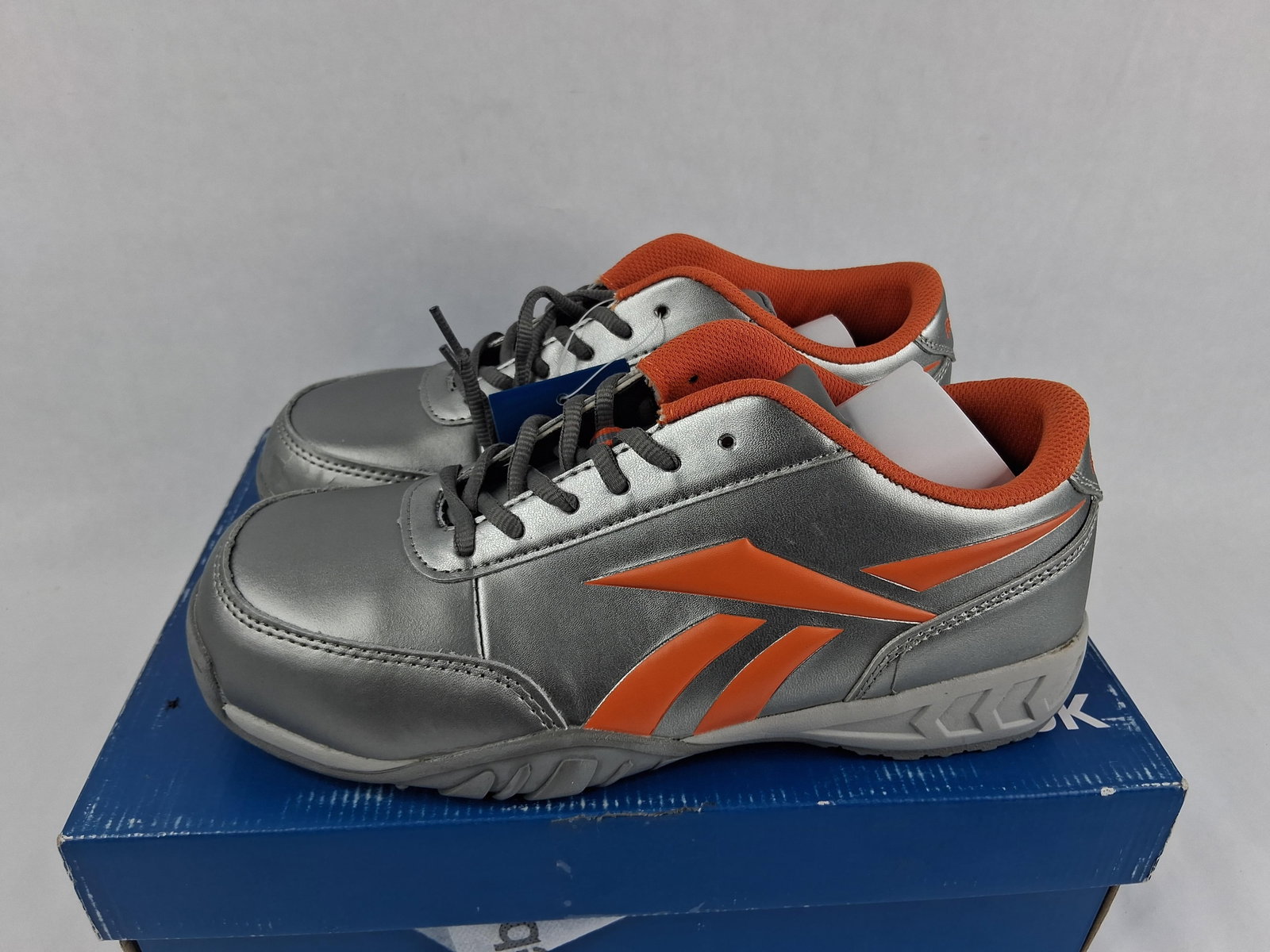 Pair Reebok Bema Women's Composite Toe Work Shoes: This pair of Reebok Work Bema Oxford safety shoes, model RB453, features synthetic metallic silver uppers with orange accents and a matching orange textile lining. The shoes are constructed with a non