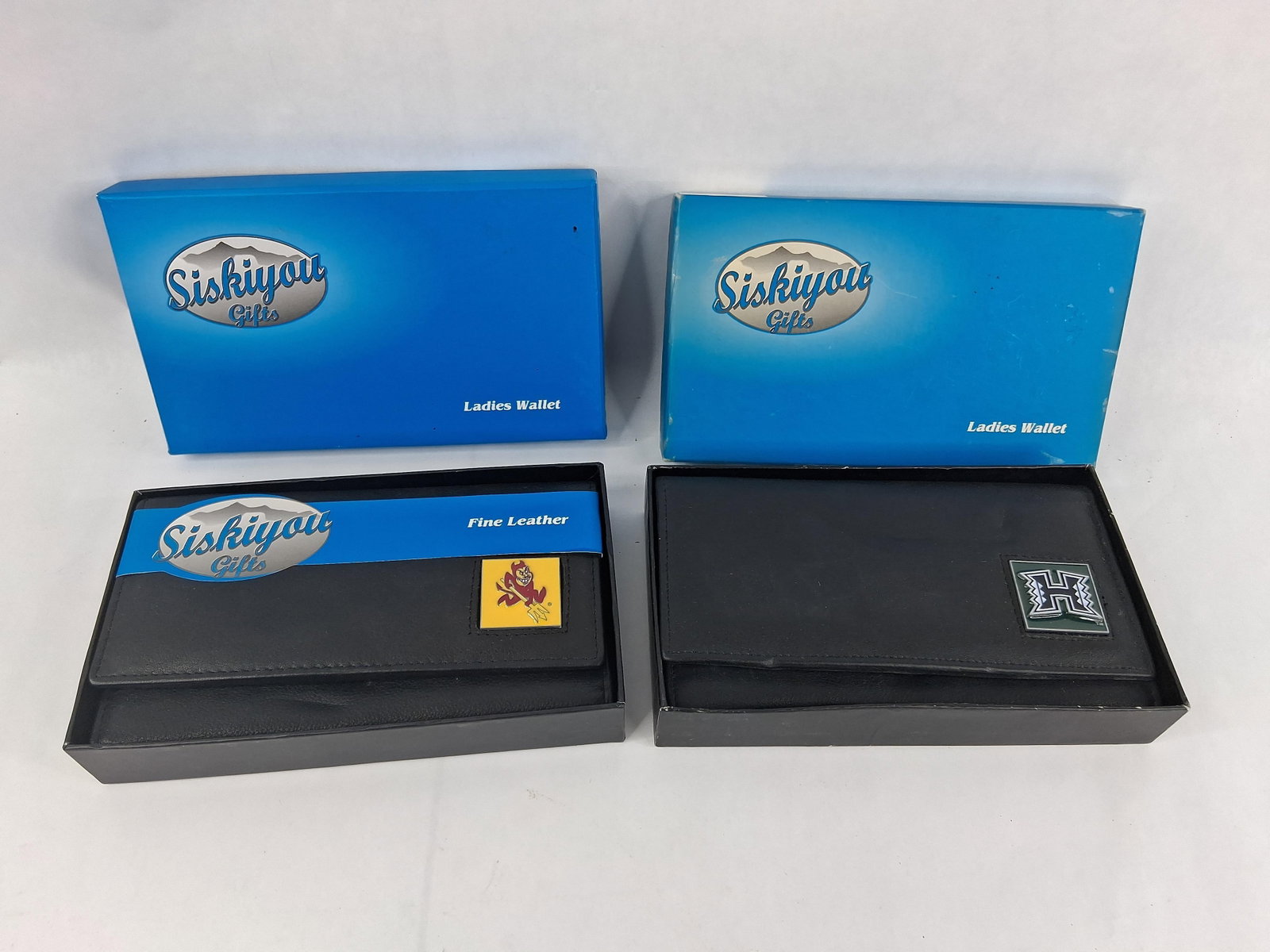 Pair Siskiyou Collegiate Leather Trifold Wallets: This lot contains two new-old-stock ladies trifold wallets manufactured by Siskiyou Gifts. Each wallet is constructed from black leather, identified as Fine Leather on the original retail packaging.