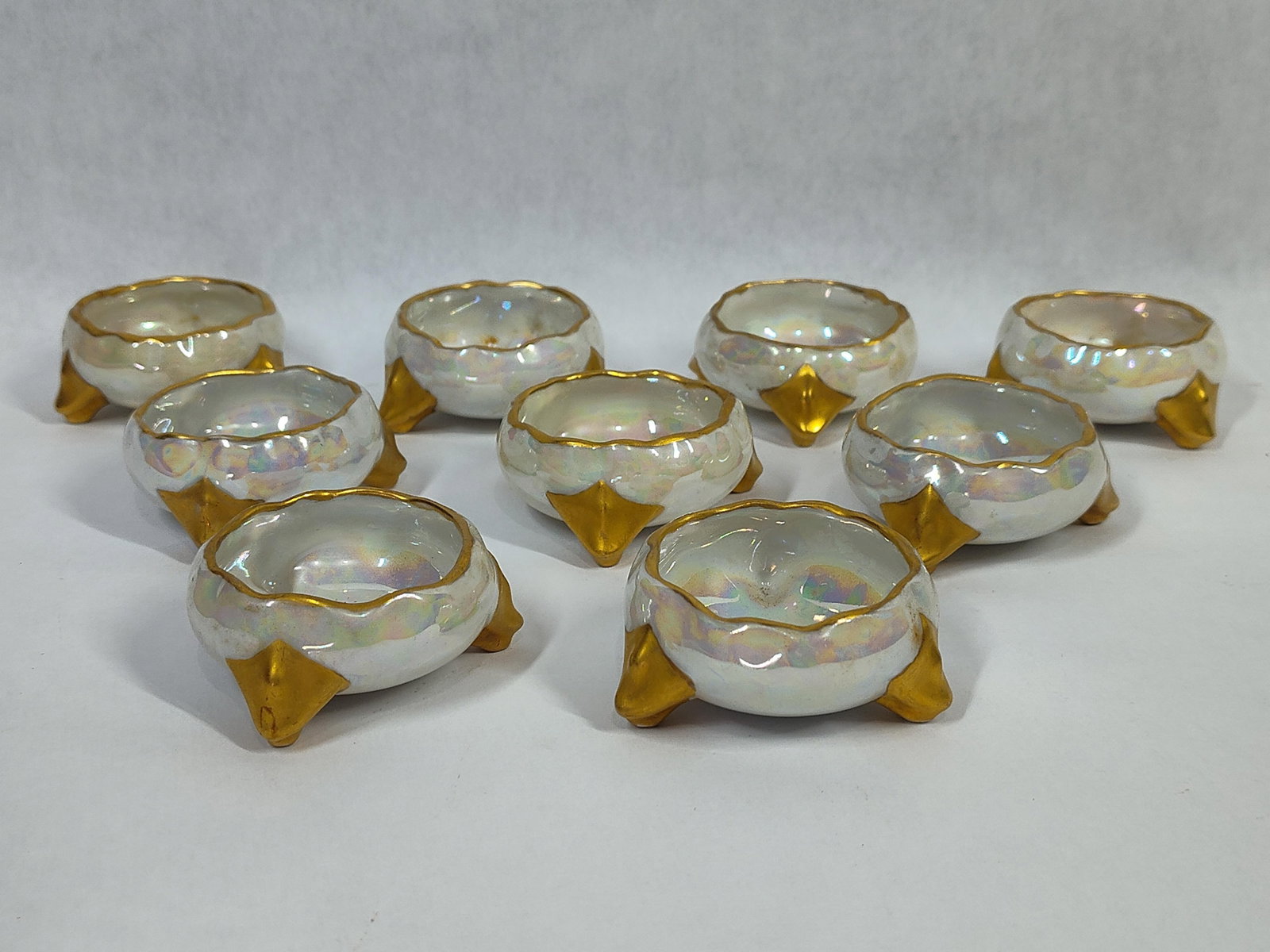 Set 9 TK Thun Iridescent Porc. Footed Salt Cellars: A set of nine porcelain salt cellars manufactured by the TK Thun factory in Czechoslovakia. Each vessel features a bulbous body with a scalloped rim, supported by three pointed tripod feet. The