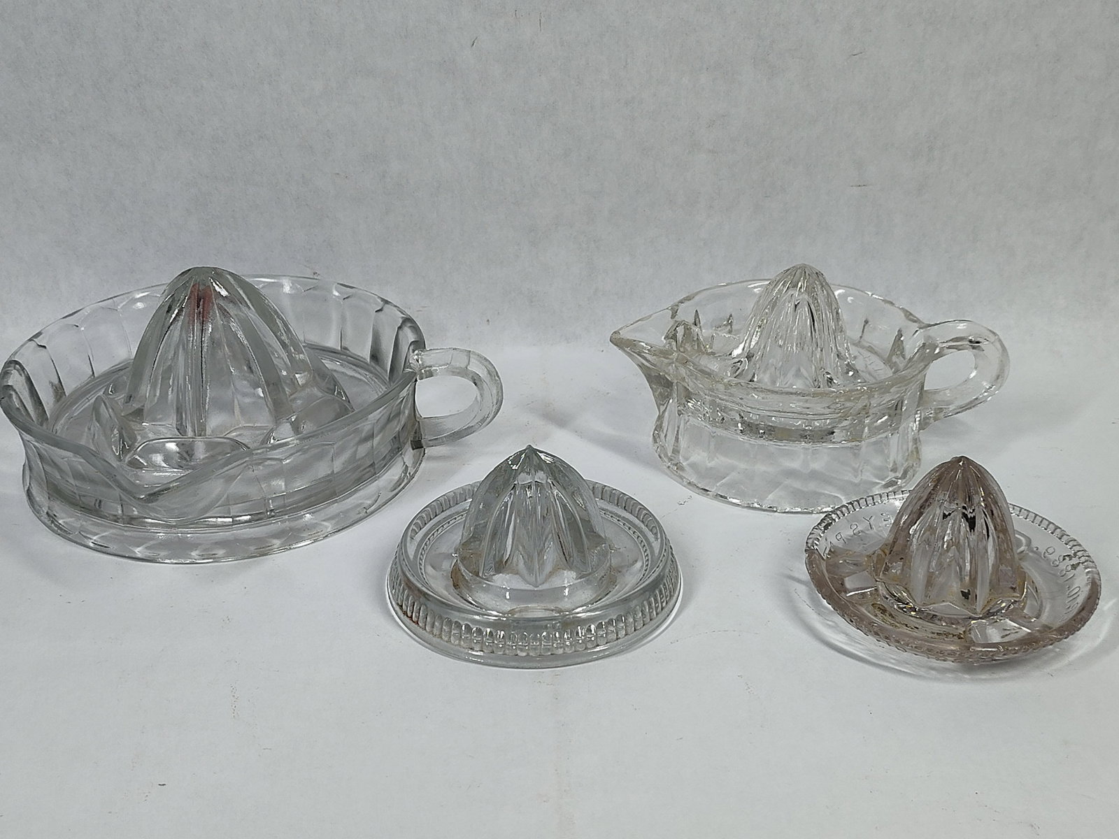 Set 4 Glass Citrus Reamers w/ 1889 Easley's Patent: A collection of four manual citrus juicers crafted in clear pressed glass, representing various designs from the late 19th through the mid-20th century. The largest reamer features a deep reservoir
