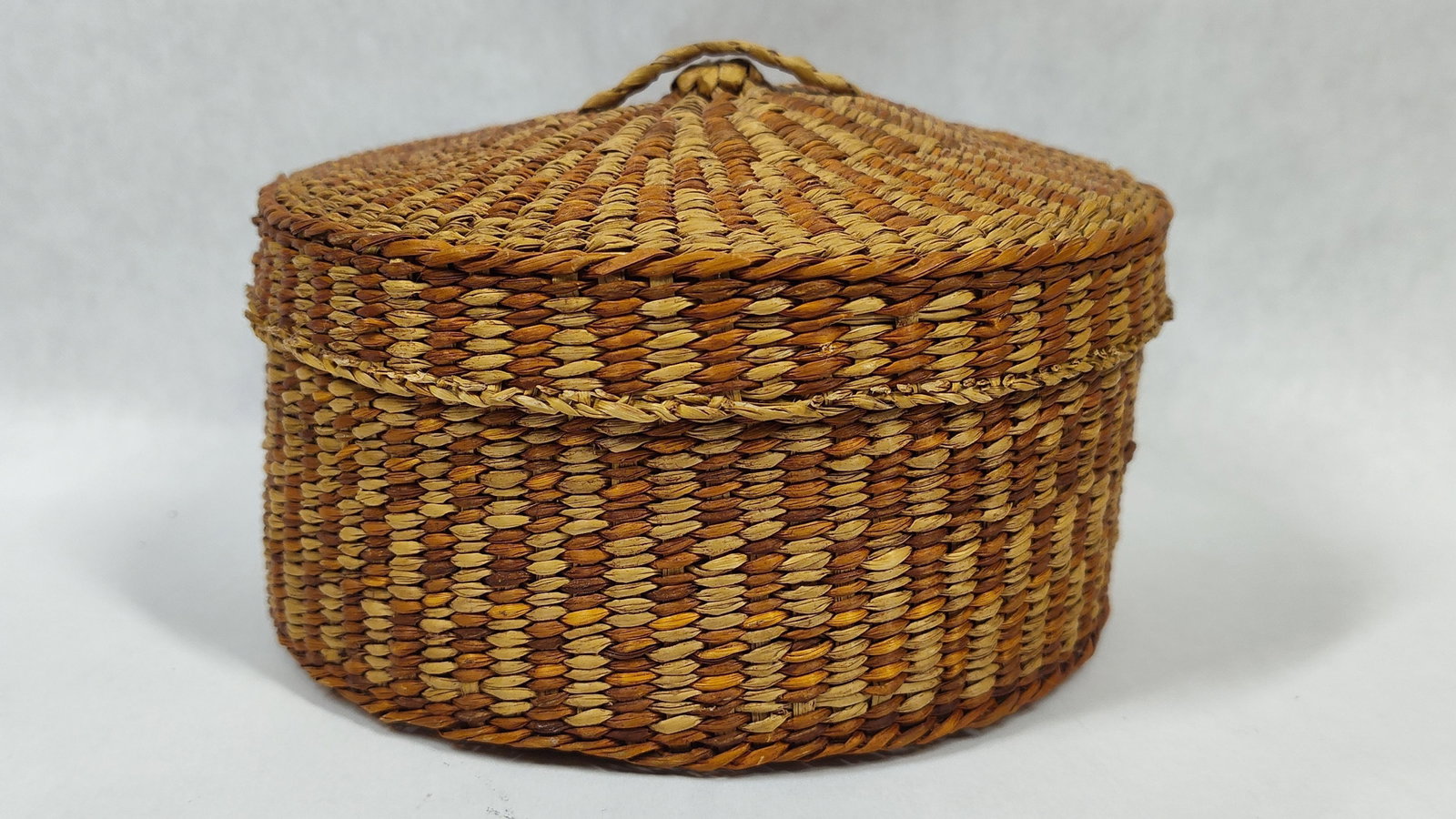 Coiled Vegetal Fiber Lidded Geometric Basket: A round lidded basket constructed from natural vegetal fibers, such as split cedar bark and dyed grasses, utilizing a tight coiled weaving technique. The exterior of the base and the lid are