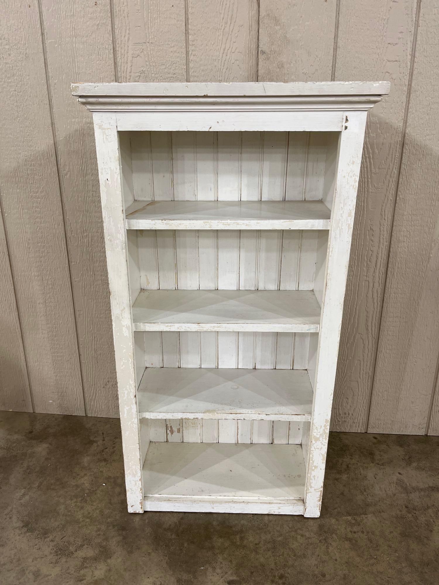 White Painted Bookcase w/ Wood Plank Back: This narrow wooden bookcase is finished in a heavily distressed white paint, characteristic of country rustic or shabby chic styling. The piece features a prominent, deep-molded crown and a simple