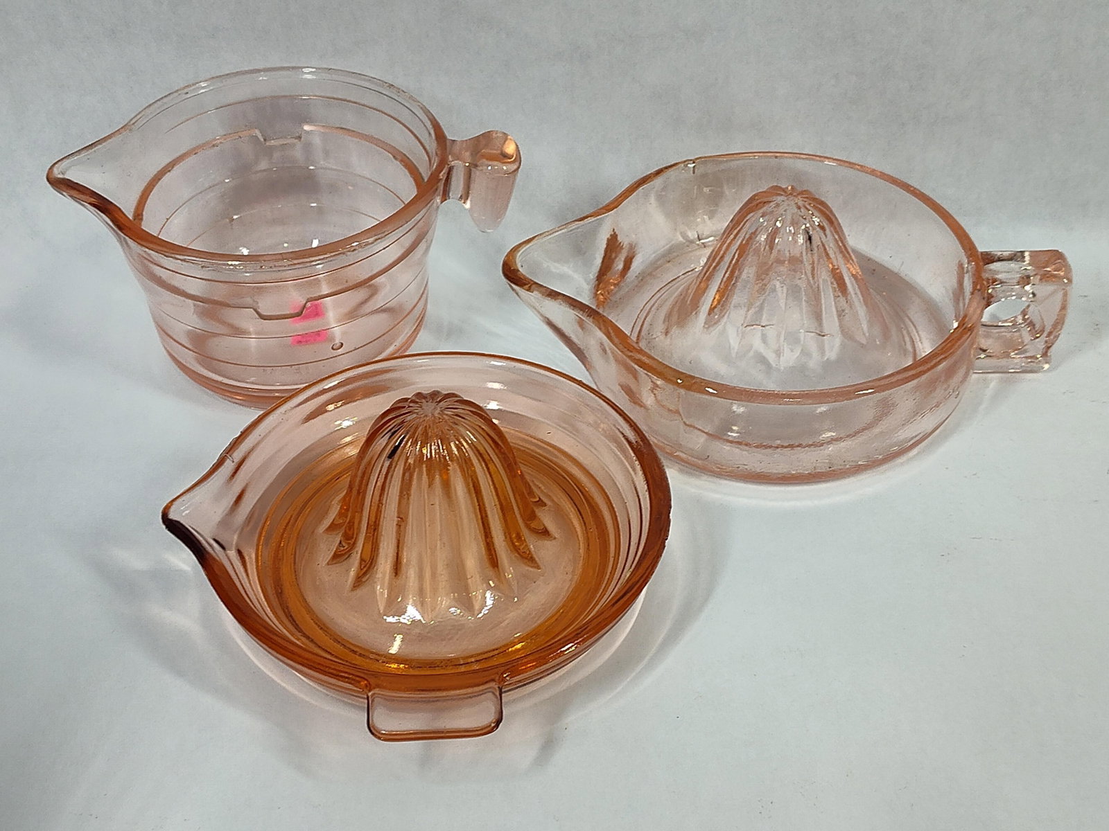 A Trio of Pink Depression Glass Citrus Juicers: A collection of three transparent pink glass kitchen items dating from the Depression era. The set includes a measuring cup base with an exterior pattern of horizontal ribbing, a single D-shaped