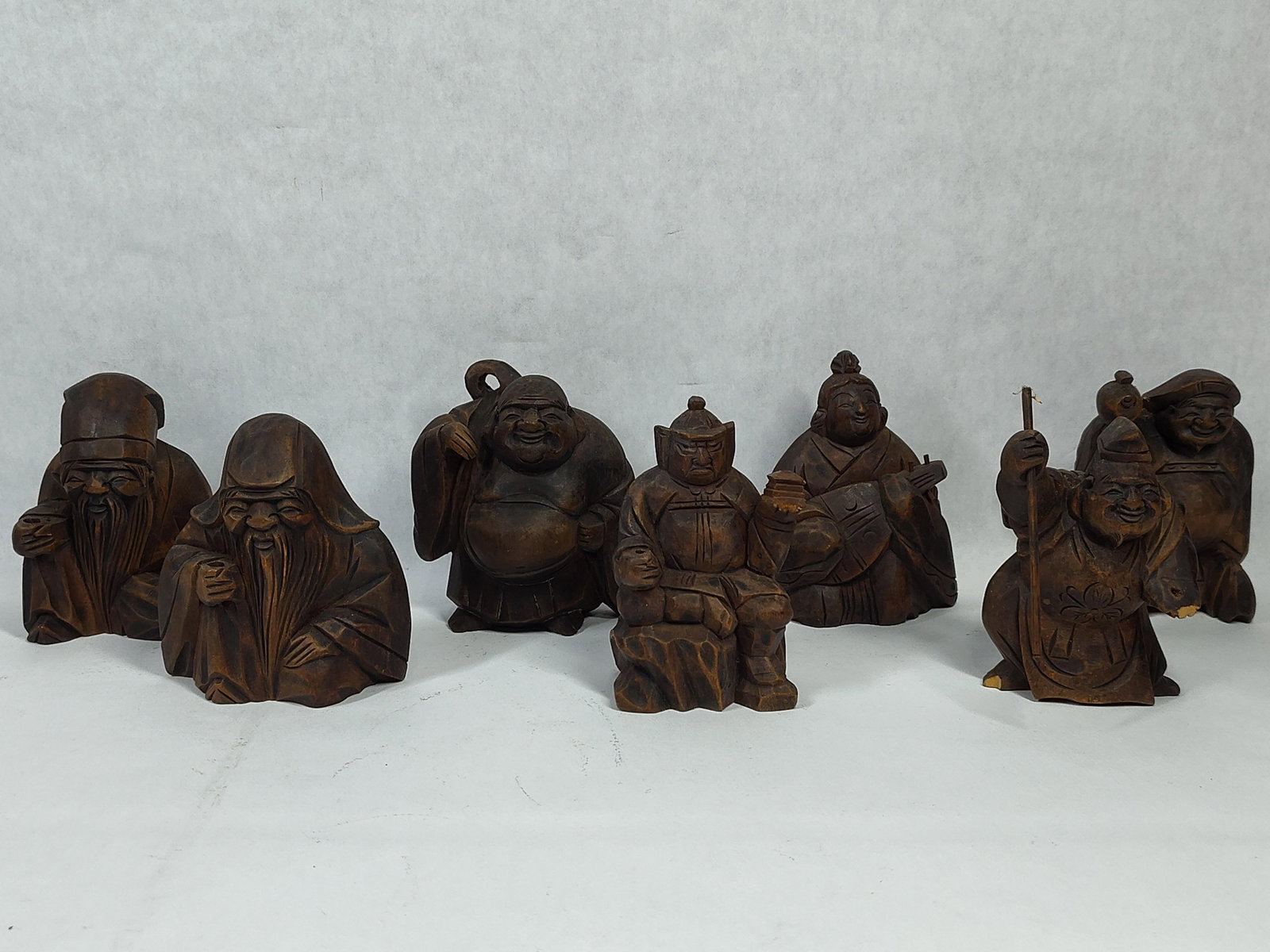 Set 7 Japanese Carved Wood Shichifukujin Figures: This collection depicts the Shichifukujin, the Seven Lucky Gods of Japanese mythology. Each figure is hand-carved from wood and finished with a dark brown stain. The set includes Hotei, characterized