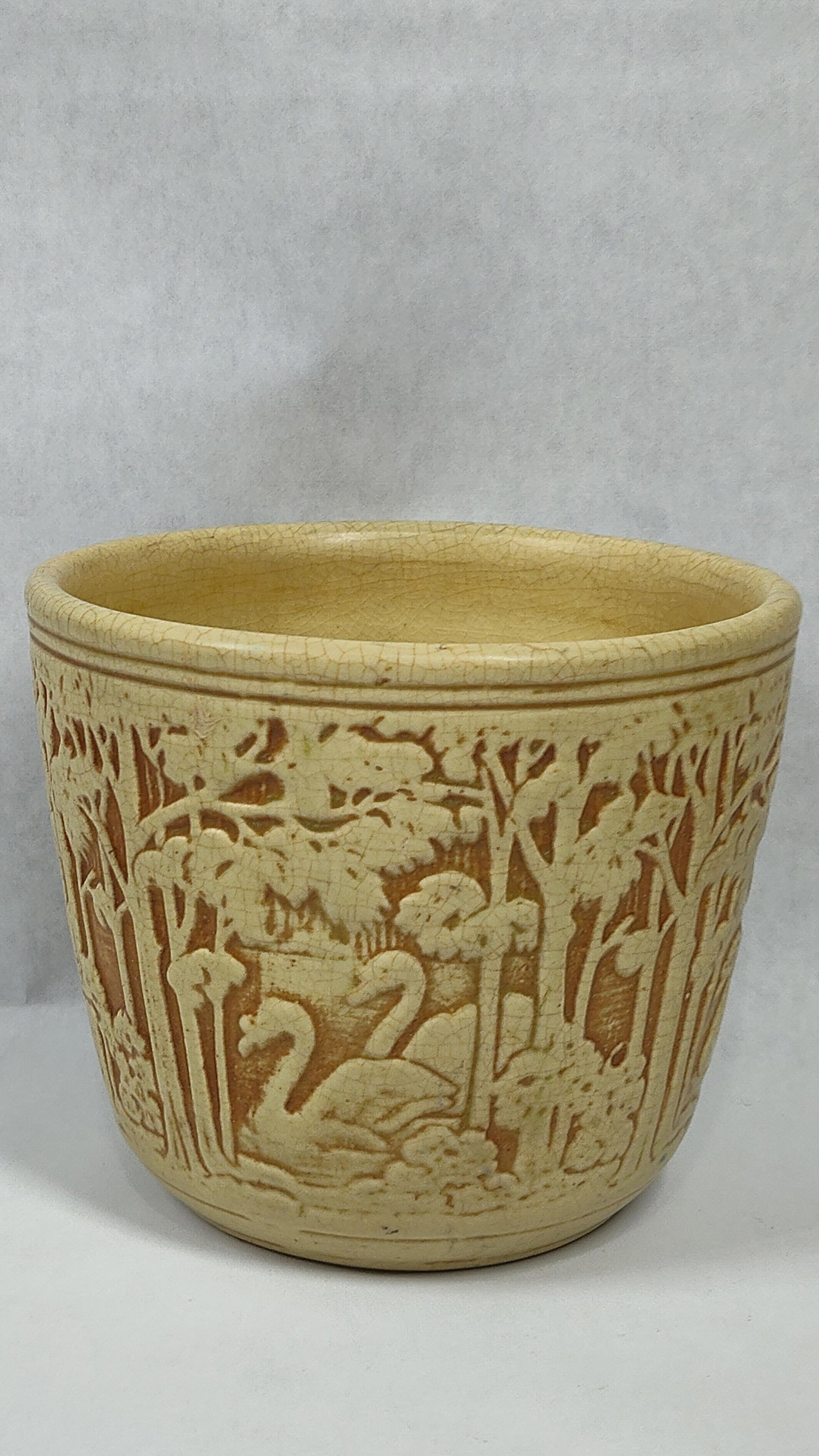 Weller Clinton Ivory Forest Swan Jardiniere: This cylindrical earthenware jardiniere is a product of Weller Pottery, Zanesville, Ohio, featuring the Clinton Ivory pattern. The exterior displays a continuous relief-molded woodland scene, with a p