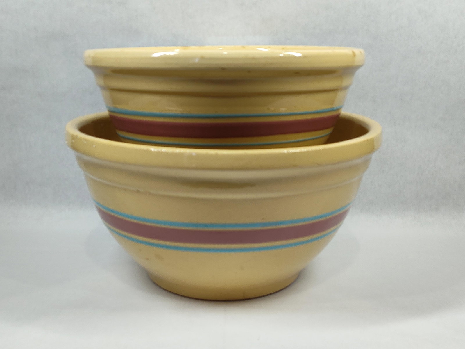 Pair Watt Pottery Banded Yellowware Mixing Bowls: A pair of yellowware ceramic nesting mixing bowls produced by Watt Pottery. Each bowl is constructed with a round form featuring a flared rim and an unglazed foot ring. The exterior of each piece is