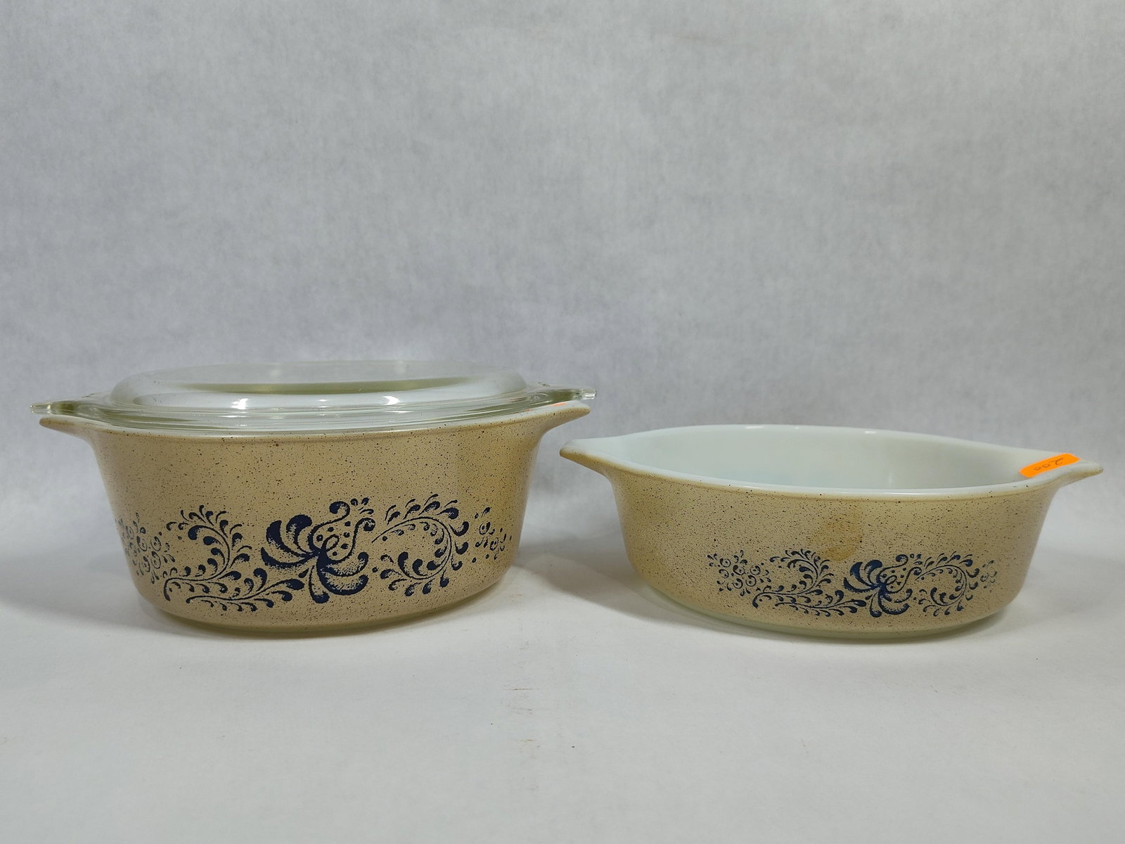A Pair of Pyrex Homestead Pattern Casserole Dishes: These two Pyrex casserole dishes feature the Homestead pattern, characterized by a speckled tan exterior finish decorated with a navy blue floral and scroll design. The vessels are constructed from op