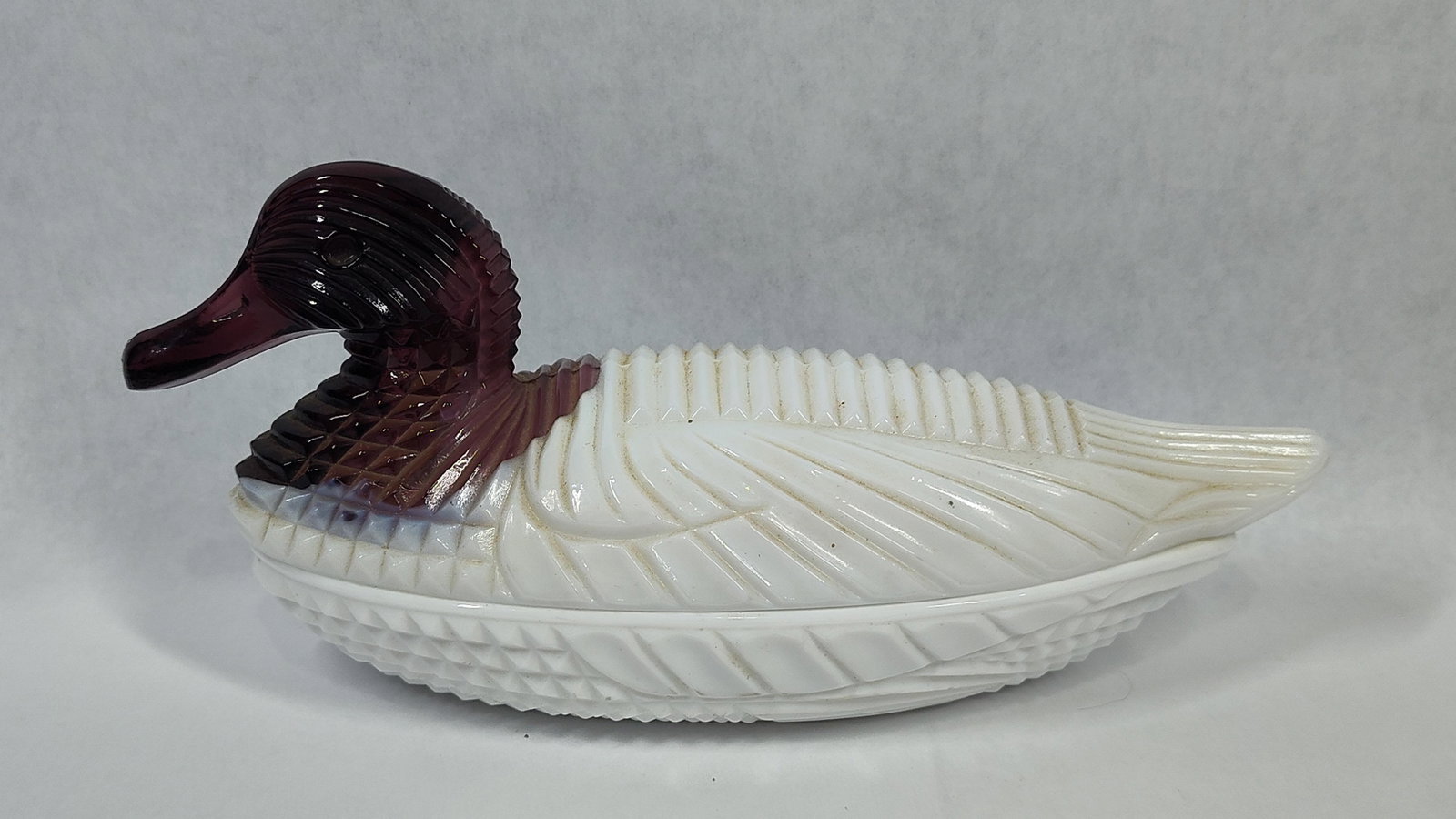 Amethyst and Milk Glass Covered Duck on Nest Dish, Attributed to L.G. Wright: A two-piece pressed glass novelty dish in the form of a duck on a nest. The lid features a translucent amethyst glass head and neck that transitions into an opaque white milk glass body. This bicolor