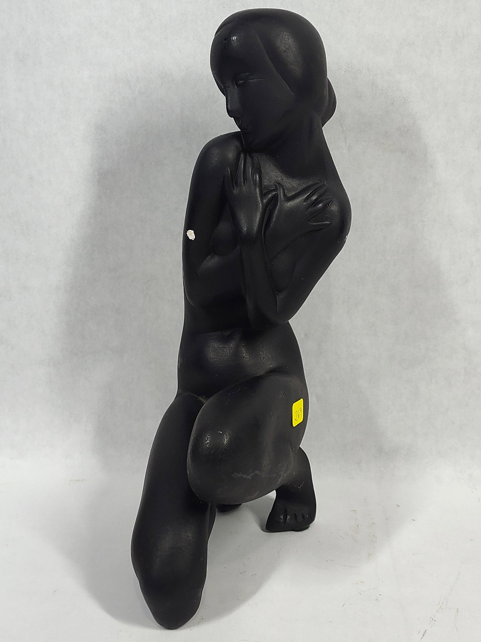 Art Deco Black Ceramic Kneeling Nude Sculpture: This Art Deco style sculpture depicts a kneeling female nude figure in a stylized pose with her arms crossed over her chest. The work is characterized by the simplified, elongated forms and smooth
