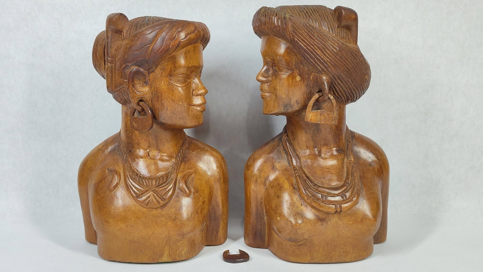 A Pair of Hand-Carved Hardwood Female Busts: A pair of hand-carved hardwood busts depicting stylized female figures. The carvings are executed in a warm-toned wood and are designed as a facing pair. Each figure displays detailed hair or head