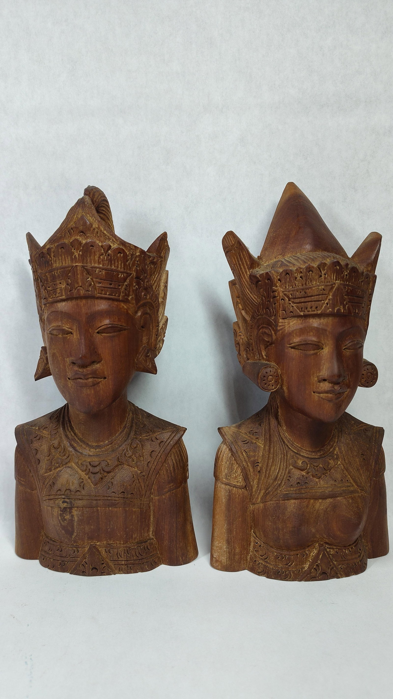 Pair Mid-20th C. Balinese Hand-Carved Wood Busts: A matching pair of hand-carved wooden sculptural busts from Bali, Indonesia, depicting a male and female figure in traditional ceremonial attire. The sculptures are crafted from wood with a warm,