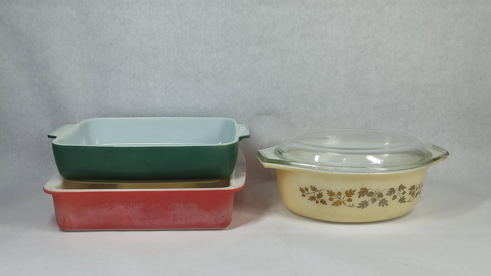 Three Vintage Pyrex Opal Glass Baking Dishes: This collection consists of three vintage Pyrex opal glass baking dishes in varied colors and forms. The first piece is a 1.5-quart oval casserole dish in the Woodland pattern, featuring a brown and t