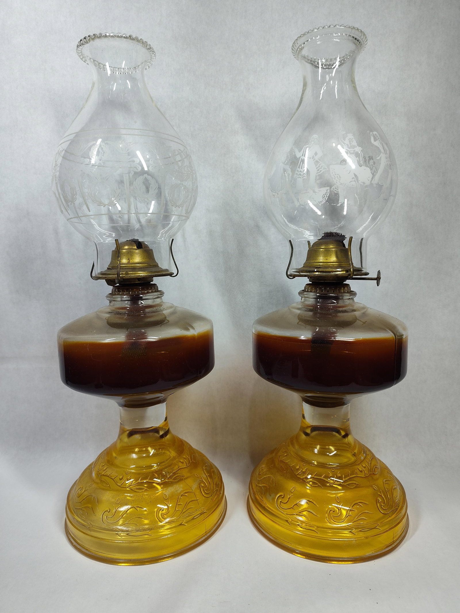 Pair Amber Glass Kerosene Lamps w/ Etched Chimneys: A pair of vintage non-electric kerosene or oil lamps featuring molded amber glass bases. The bases are decorated with embossed scroll and foliate patterns and support clear glass reservoirs. The