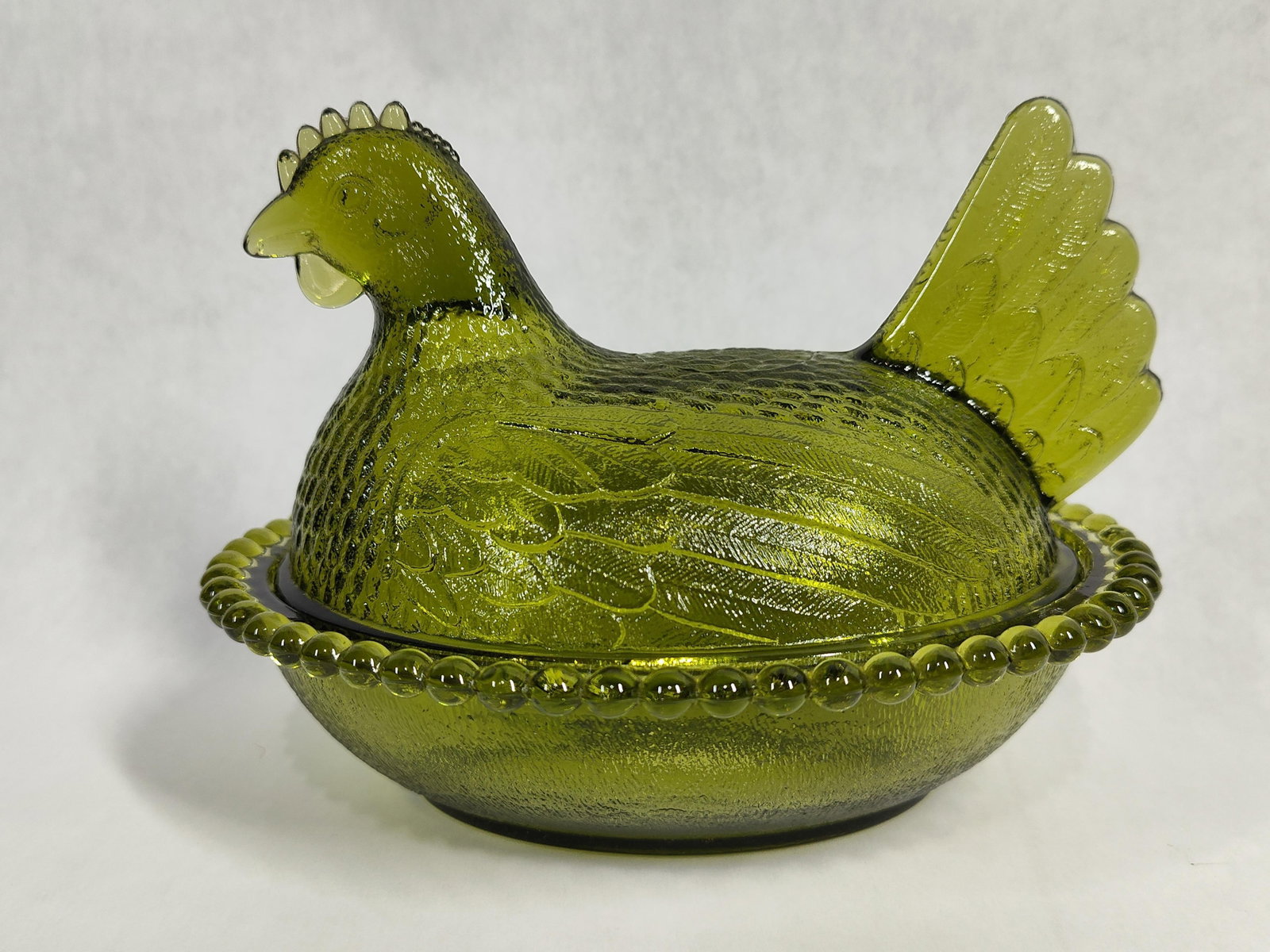 Olive Green Hen on Nest Pressed Glass Covered Dish: A two-piece covered dish in the form of a nesting hen, attributed to the Indiana Glass Company. The item is manufactured from translucent pressed glass in a deep olive green color. The upper lid
