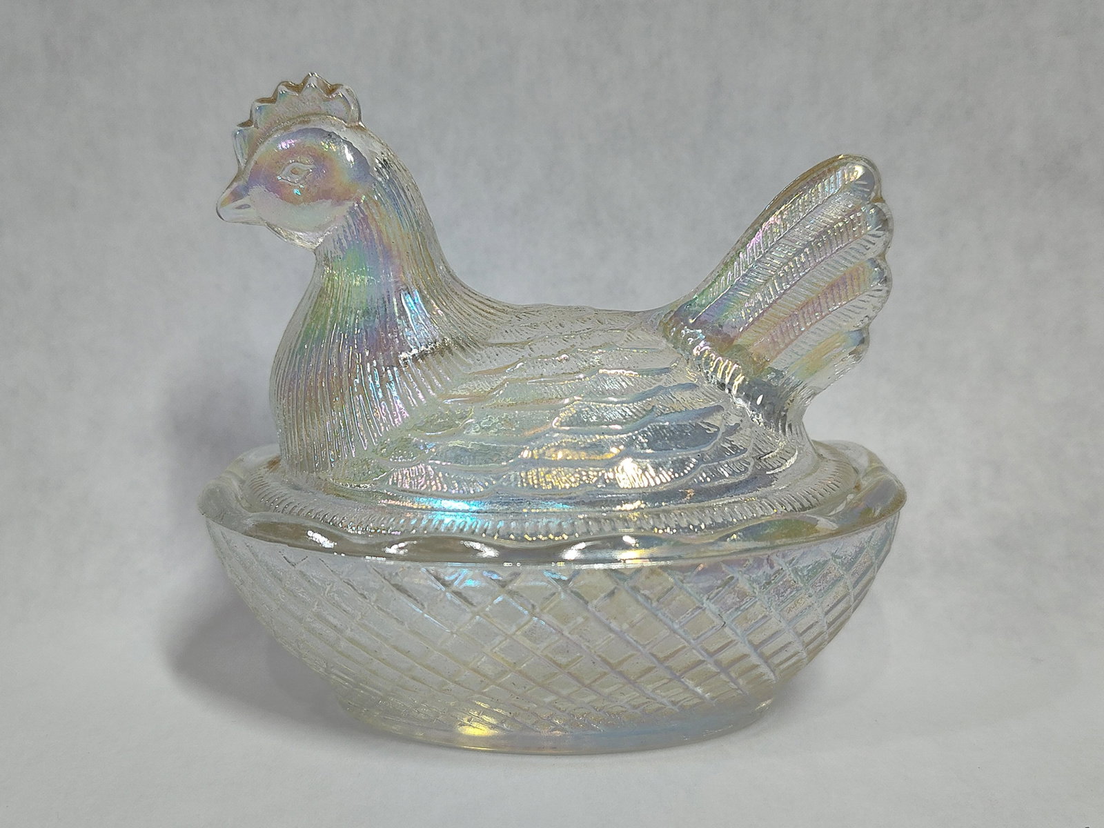 L.E. Smith Iridescent Hen on Nest Covered Dish: This is an iridescent clear glass covered dish in the classic hen on nest form. The object consists of a molded lid in the shape of a hen with detailed feathering and an oval base with a