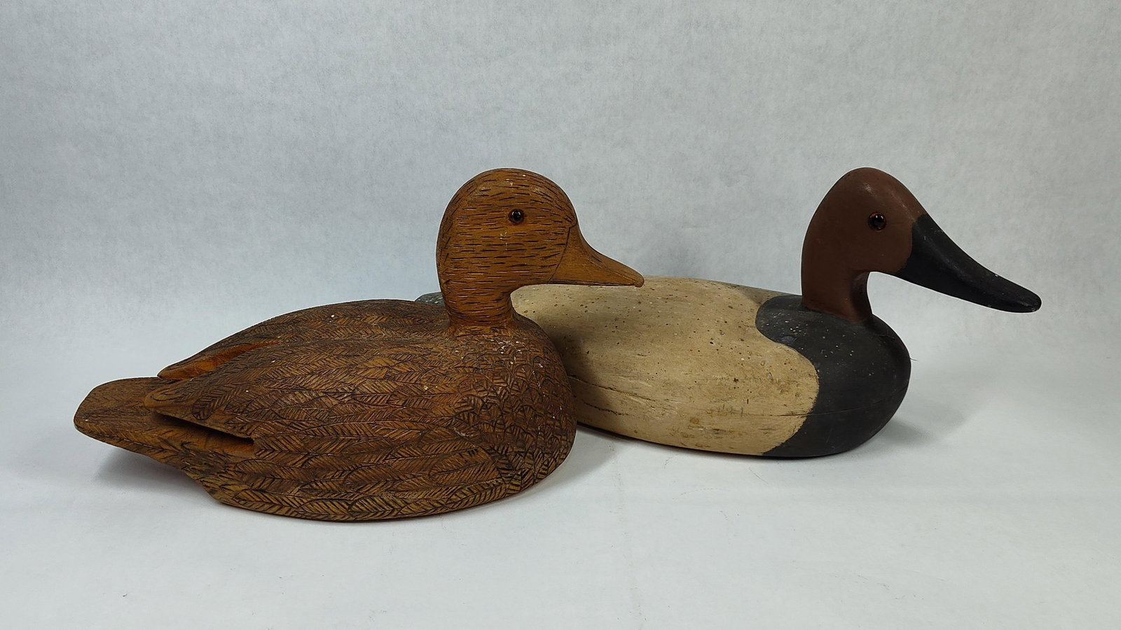 Paul Dziak Pair Hand-Carved Wood Duck Decoys: This pair of hand-carved wooden duck decoys includes one specimen with a natural finish and one specimen with a painted finish. The natural-finish decoy features detailed carved textures representing