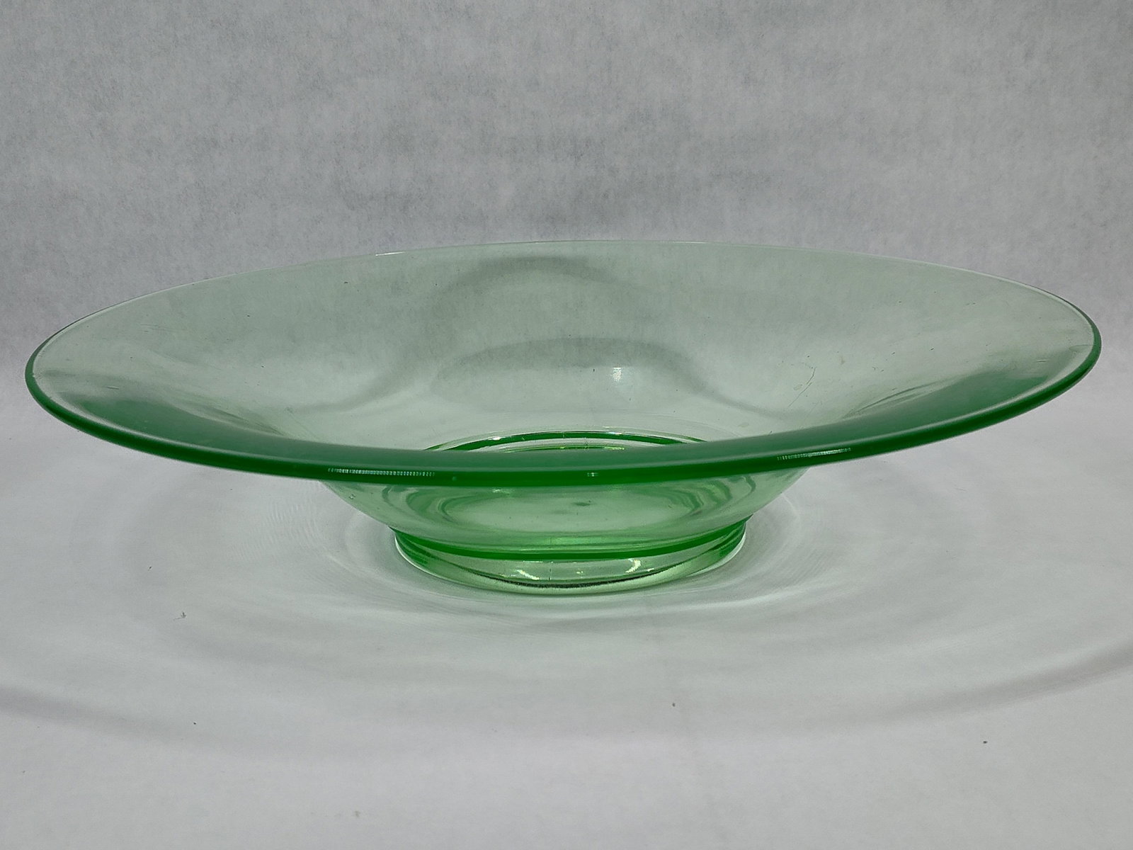 Large Green Uranium Glass Footed Console Bowl: A vintage American footed console bowl crafted from transparent light green uranium glass. The piece is designed as a centerpiece with a shallow basin and a broad, wide rim supported by a circular
