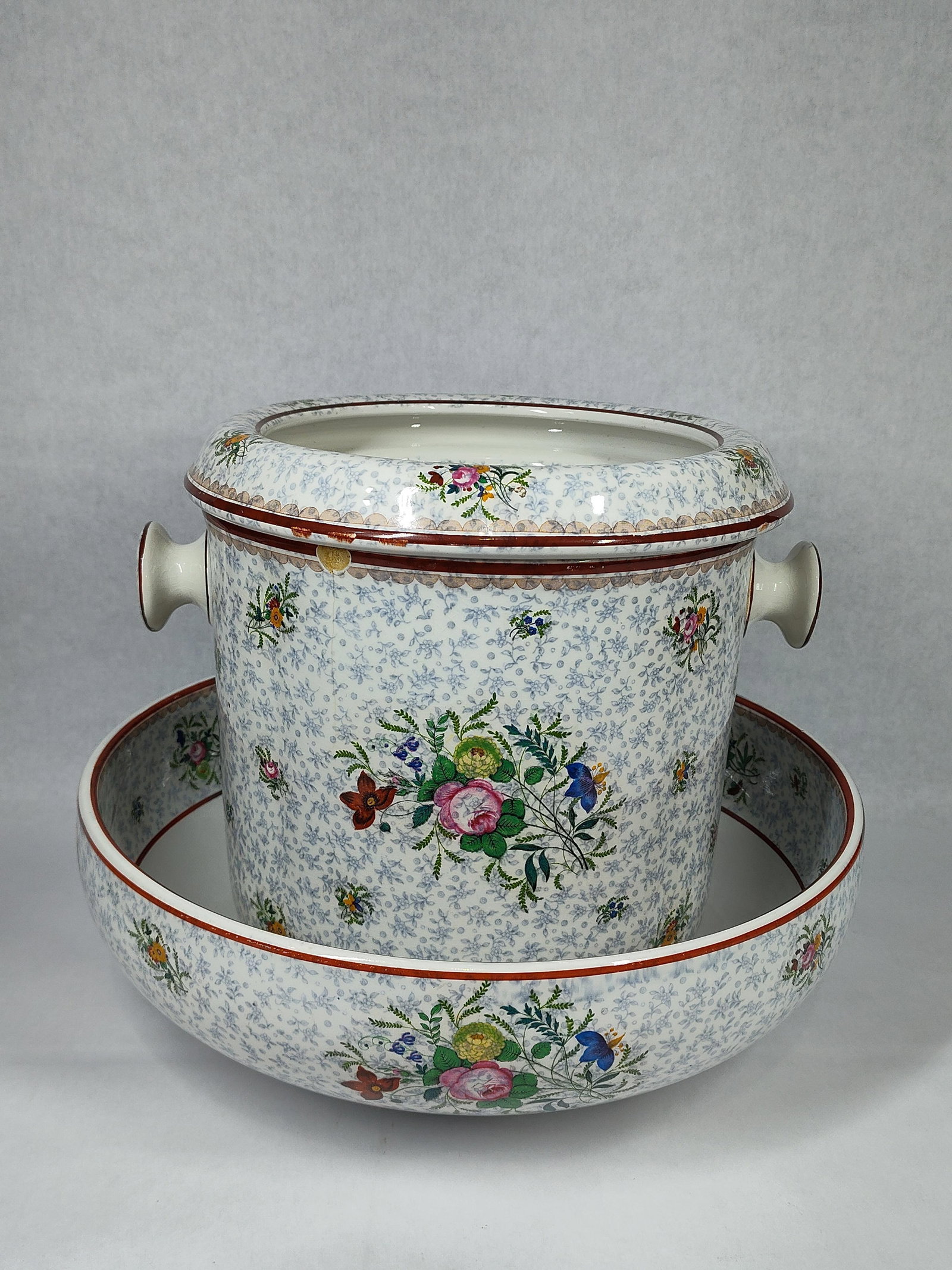 Doulton Burslem Floral Chintz Washstand Set: This ceramic washstand set includes a cylindrical slop jar with a recessed vented lid and a large circular wash basin. Both pieces are made of glazed earthenware and feature a blue transfer-printed