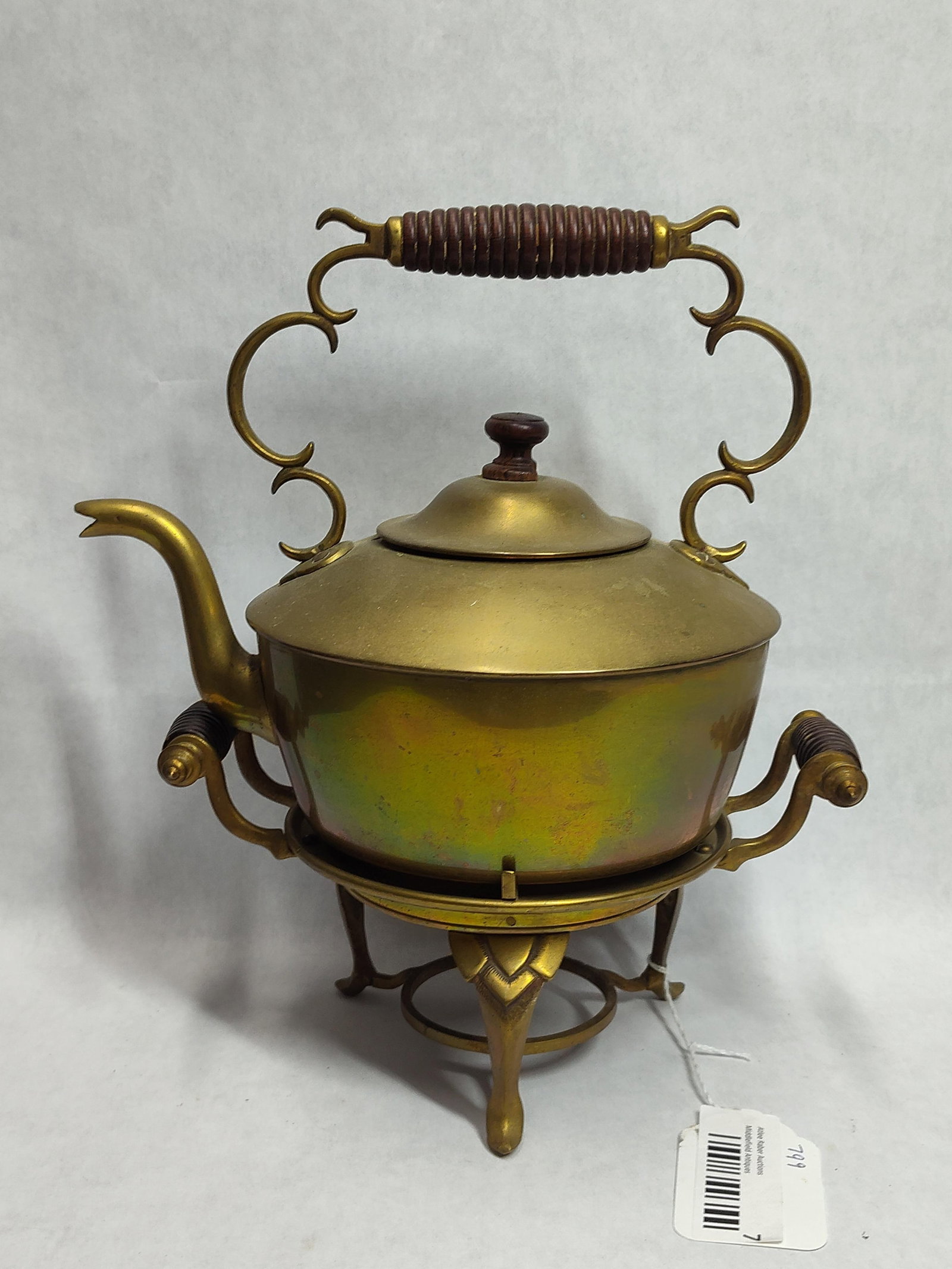 Victorian Style Brass Spirit Kettle on Stand: A decorative brass tea kettle on a matching tripod stand. The kettle features a spun brass body with a curved spout and a fitted lid topped with a wooden finial. It is equipped with a large overhead