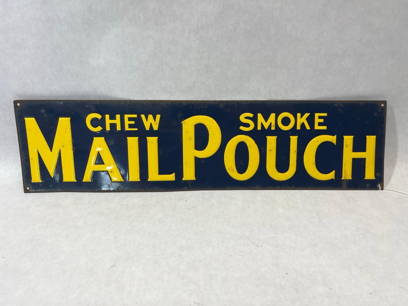 Mail Pouch Tobacco Embossed Metal Sign: A rectangular advertising sign for Mail Pouch Tobacco. The sign is made of embossed metal with a dark blue painted finish and yellow lettering. The central text features the brand name MAIL POUCH in
