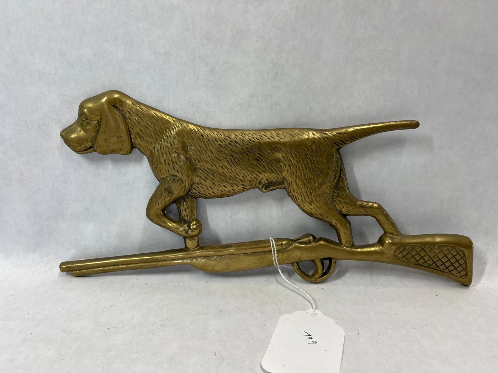 Cast Brass Hunting Dog & Shotgun Wall Plaque: A decorative wall plaque made of cast brass, depicting a hunting dog, such as a Pointer or Setter, in an active field stance above a shotgun. The dog figure is detailed with a textured fur pattern,
